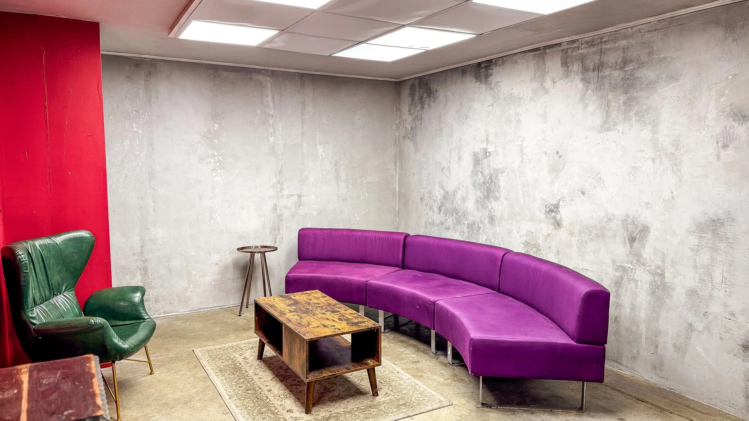 A lounge with a plain concrete wall, a curved purple sofa, a green armchair, a small wooden table, and a ceiling with fluorescent lights.