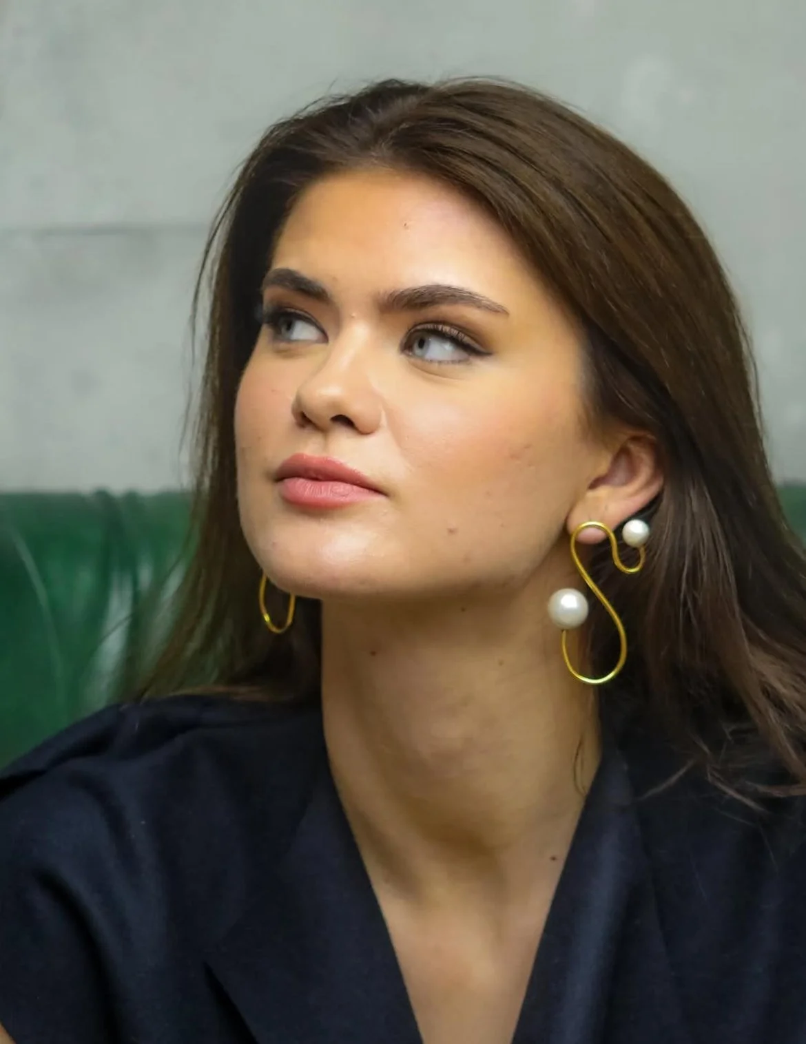 Close-up of a woman with brown hair, blue eyes, wearing gold and pearl earrings and a black top, looking to the side against a plain background.