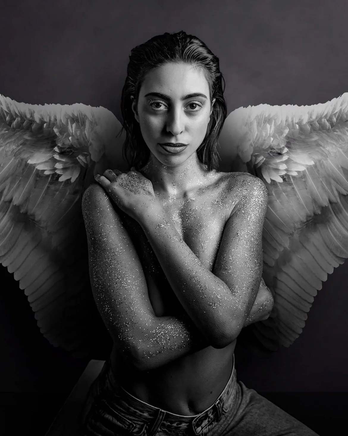 Black and white photo of a woman with wings, covered in glitter, posing with crossed arms and a confident expression.