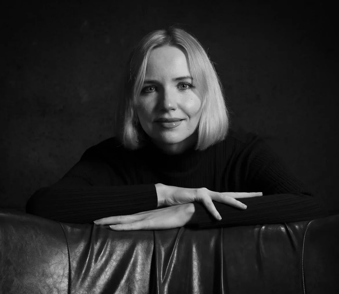 Black and white portrait of a woman with shoulder-length blonde hair, wearing a long-sleeved dark top, resting her arms on the back of a leather chair, looking at the camera with a slight smile.