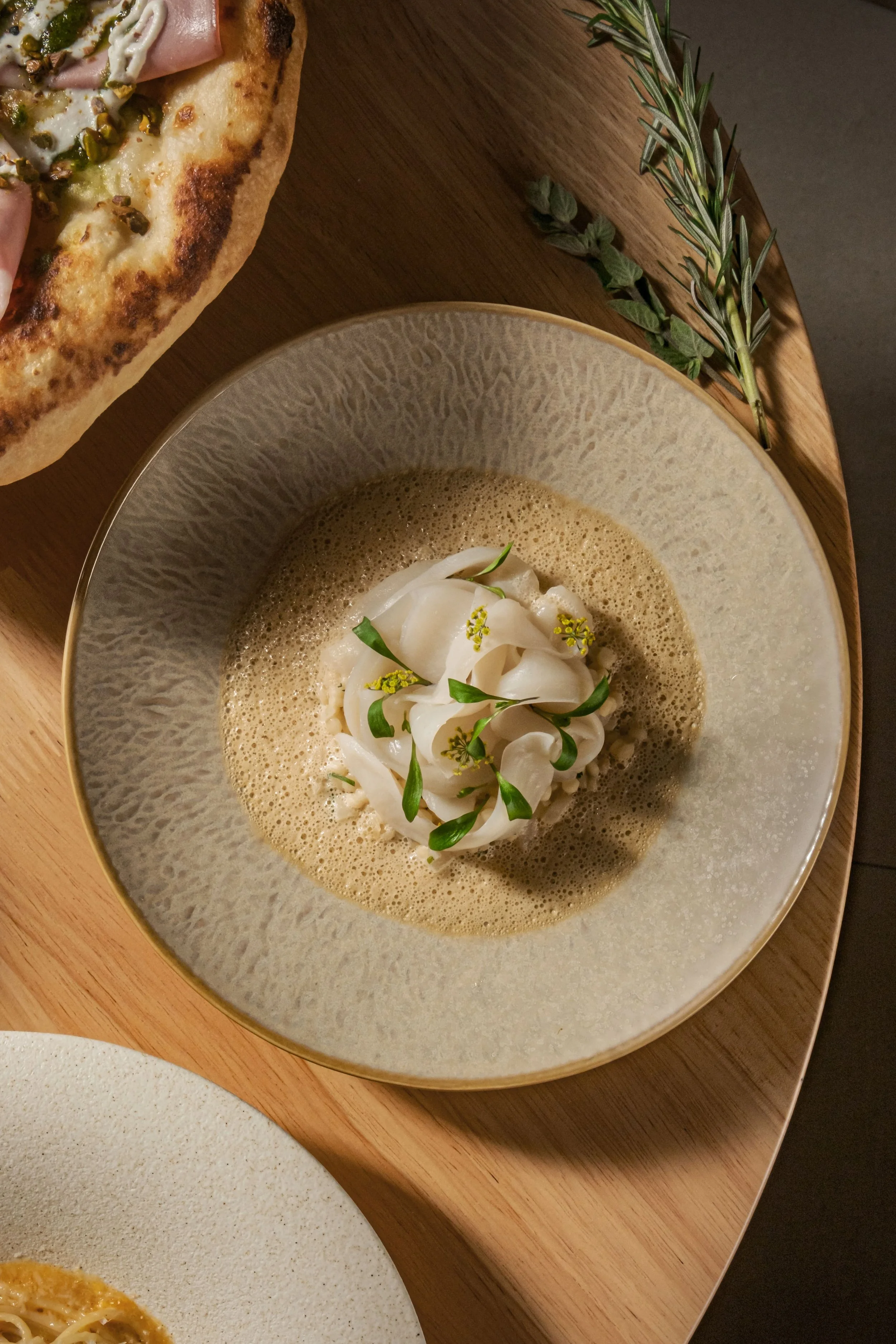 A bowl of creamy pasta topped with white sauce, small green garnishes, and flowers, placed on a wooden tray with herbs. Part of a pizza with toppings is visible in the corner.