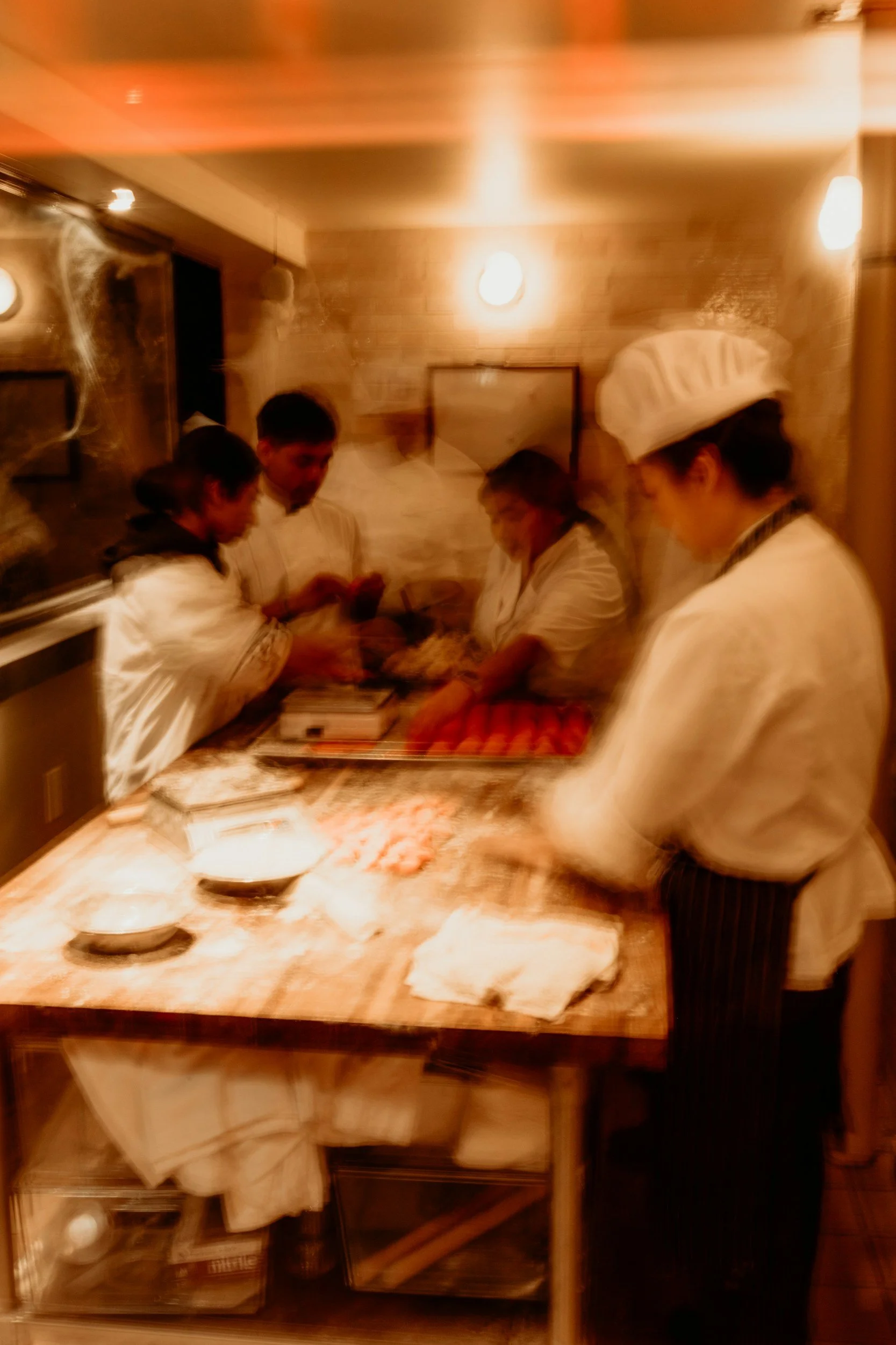 Cooks preparing food in a kitchen, with a person dressed in a chef's uniform and hat, working at a counter with bowls of ingredients and dough.