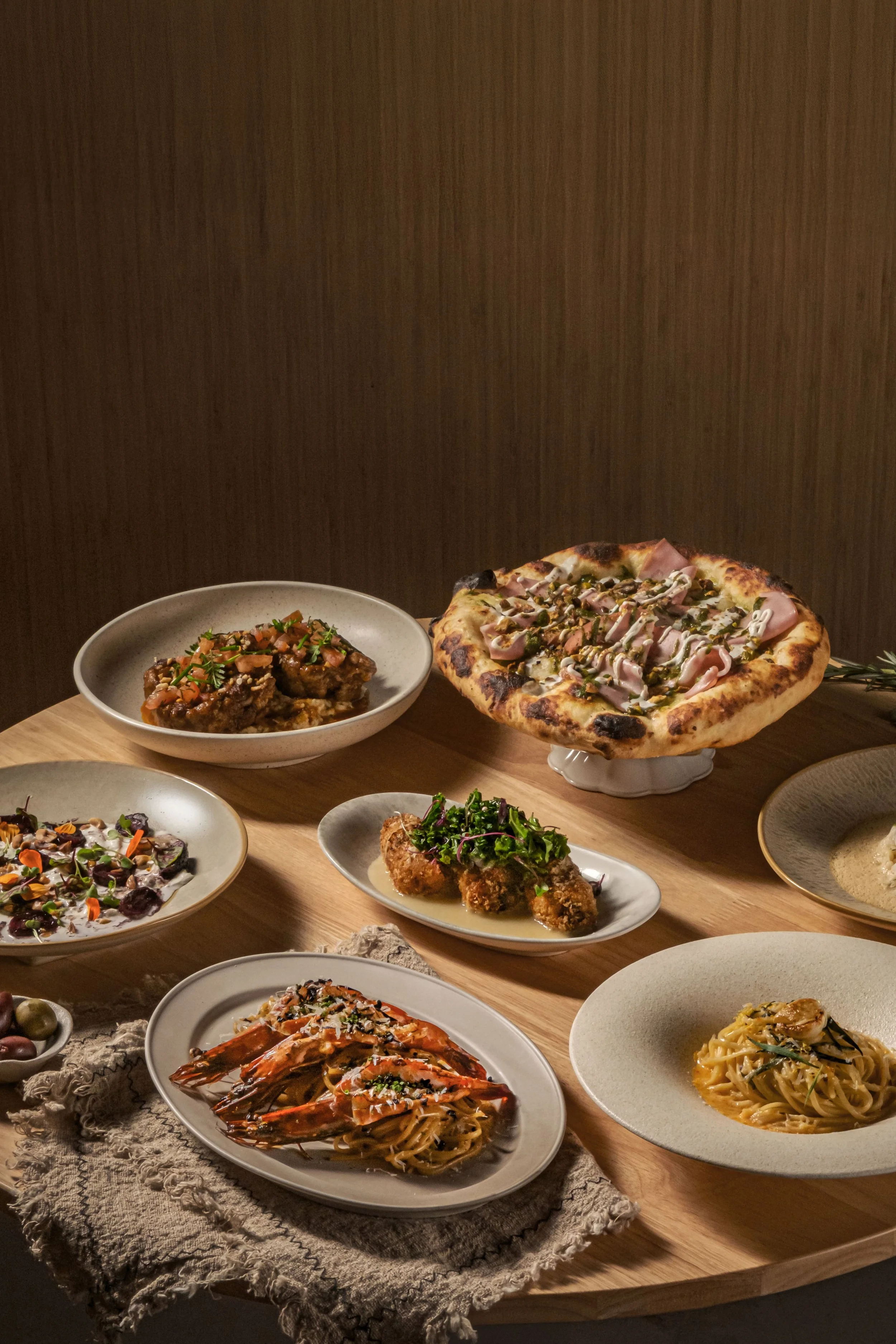 A variety of dishes on a wooden table, including a pizza with ham and peppers, a plate of spaghetti with shrimp, fried appetizers, salads, and other pasta dishes, arranged for a meal.