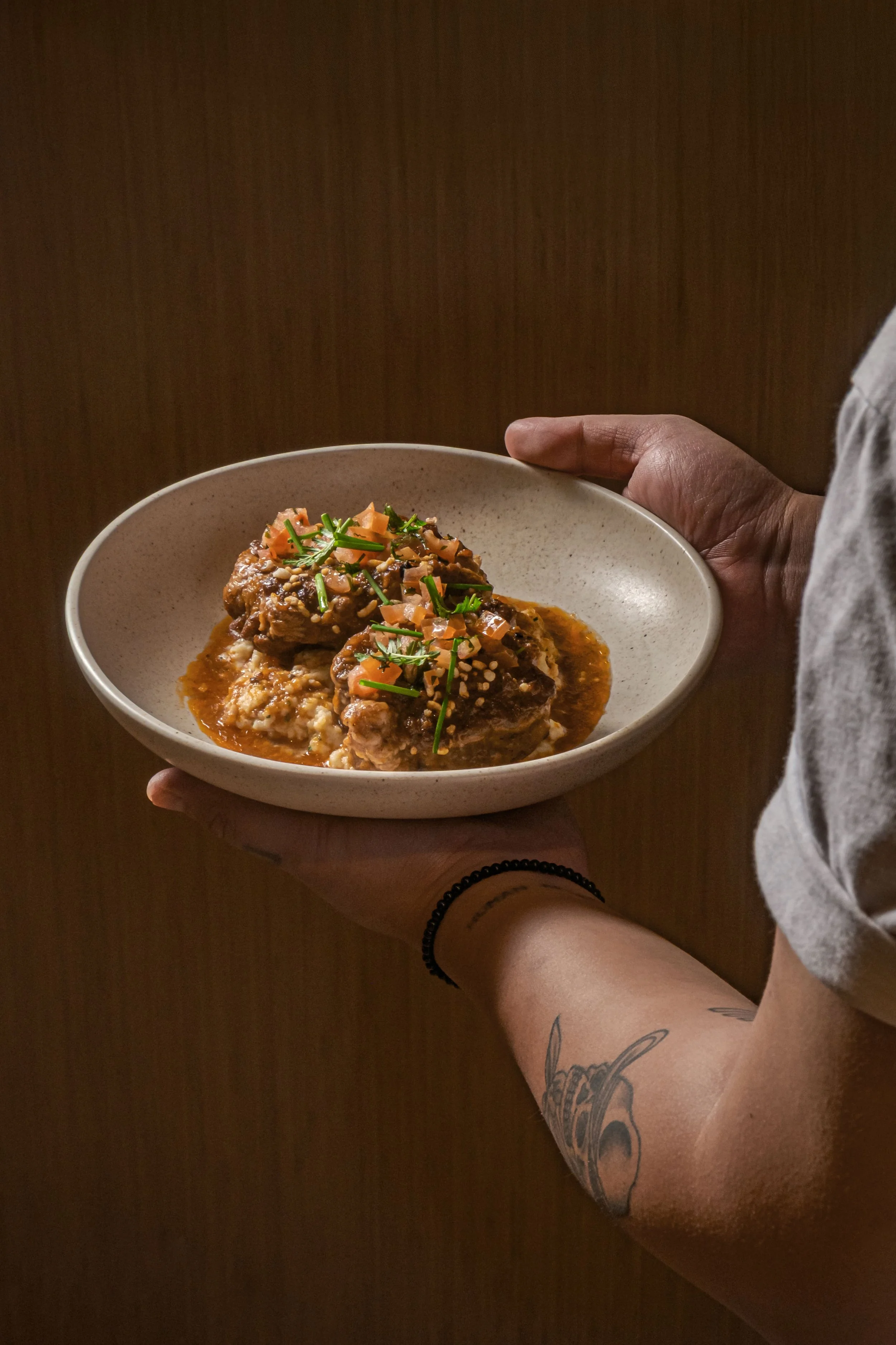 A person holding a ceramic bowl filled with cooked food, garnished with chopped herbs and vegetables, with a wooden background.