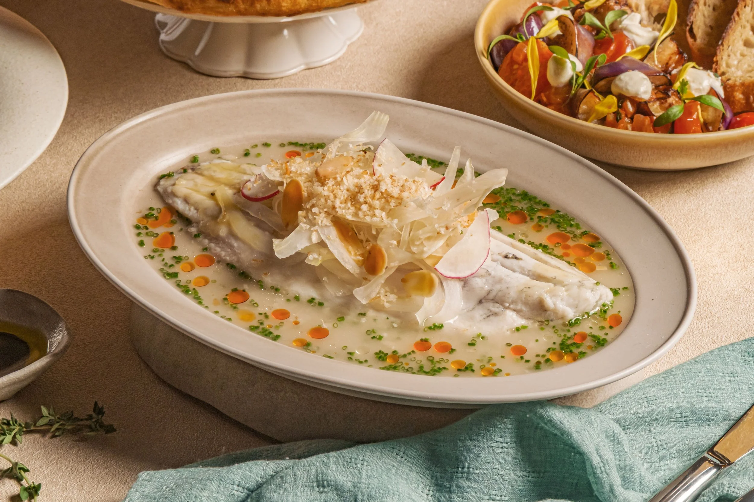 A plate of cooked white fish garnished with sliced radishes, almonds, and a creamy sauce, surrounded by bowls of salad, bread, and dipping sauces.