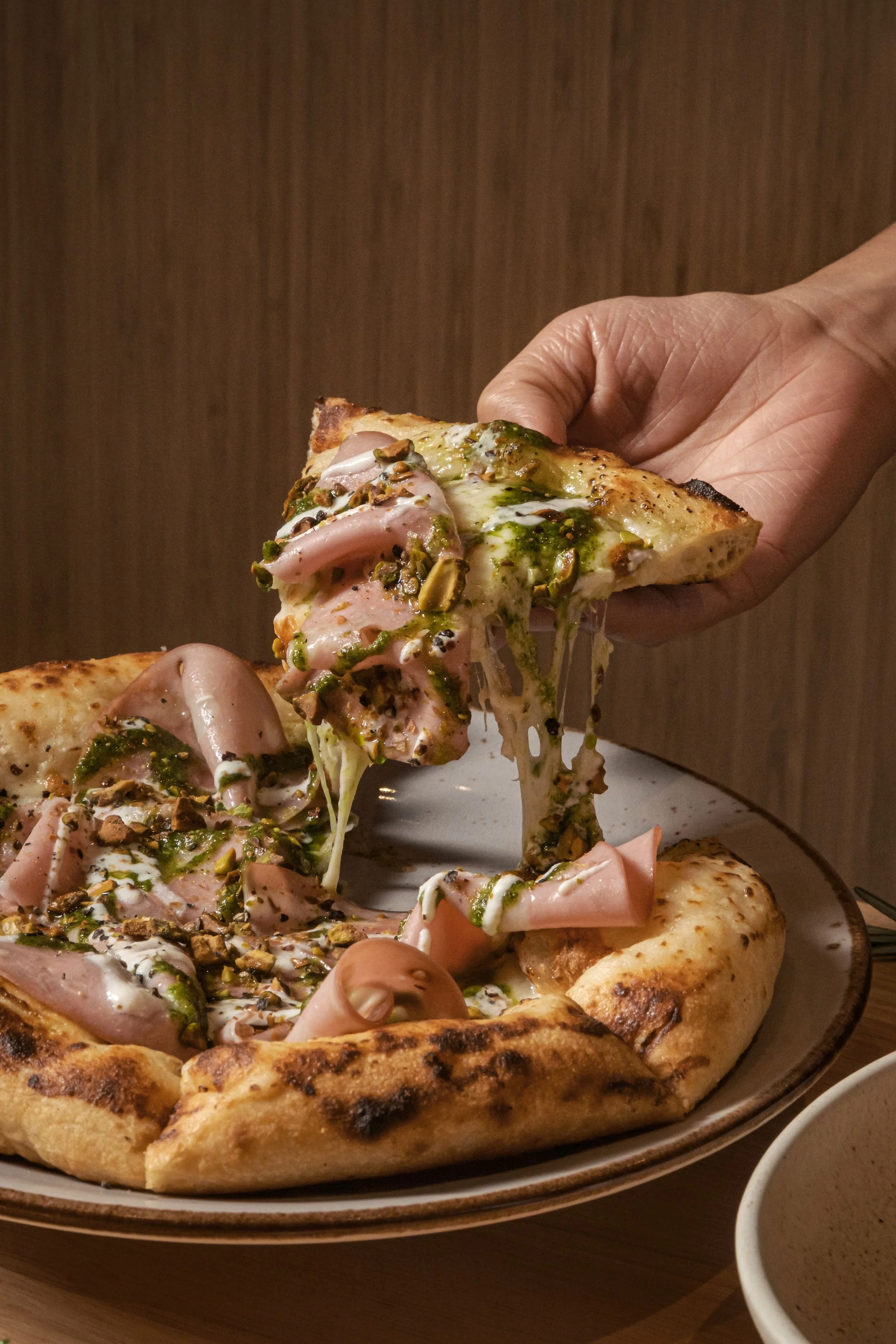 A slice of pizza being lifted from a plate, topped with ham, melted cheese, green sauce, and chopped nuts, with a wooden textured background.