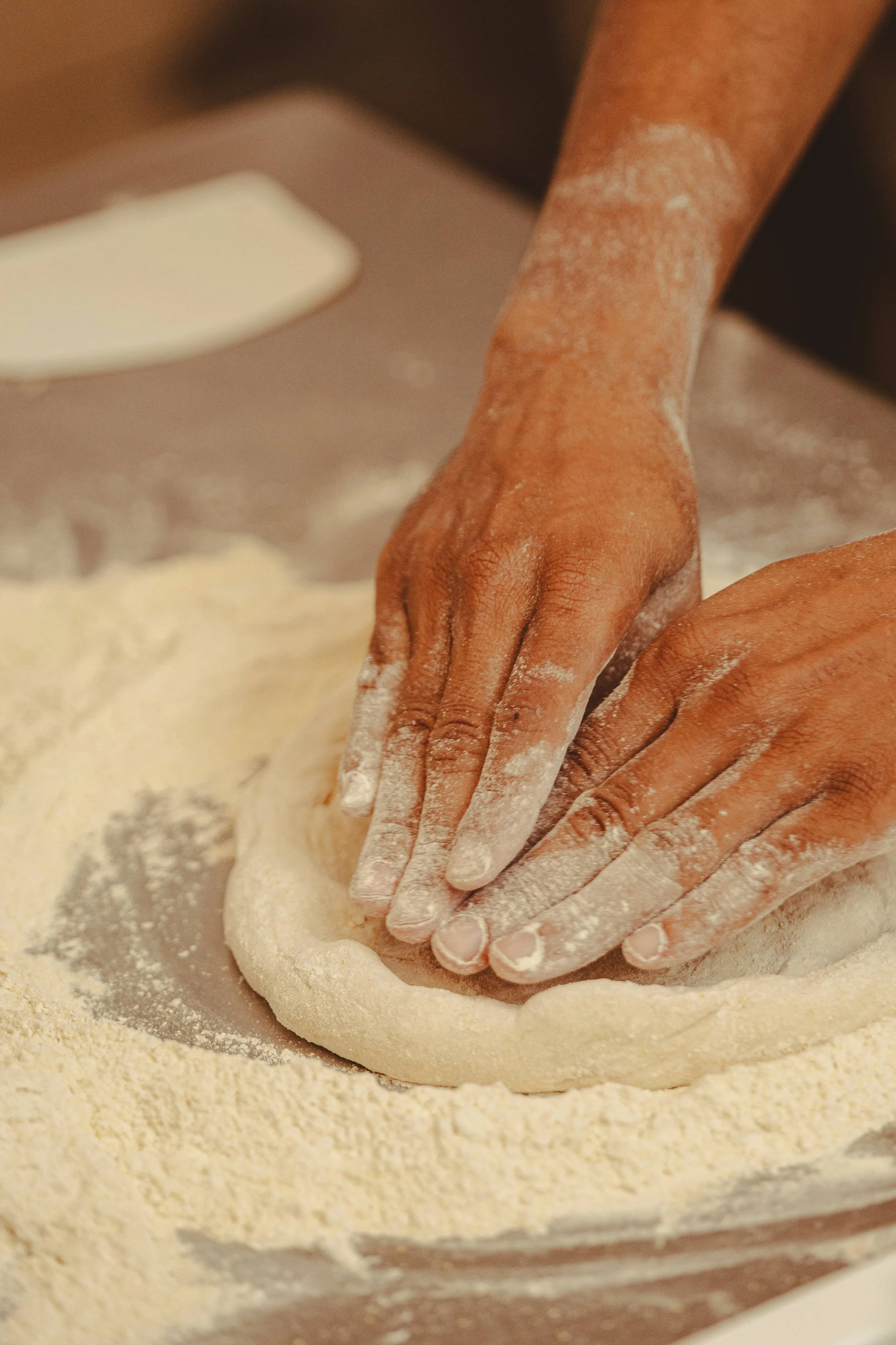 Close-up of hands pressing down on pizza dough dusted with flour, with a white bowl partially visible in the background.