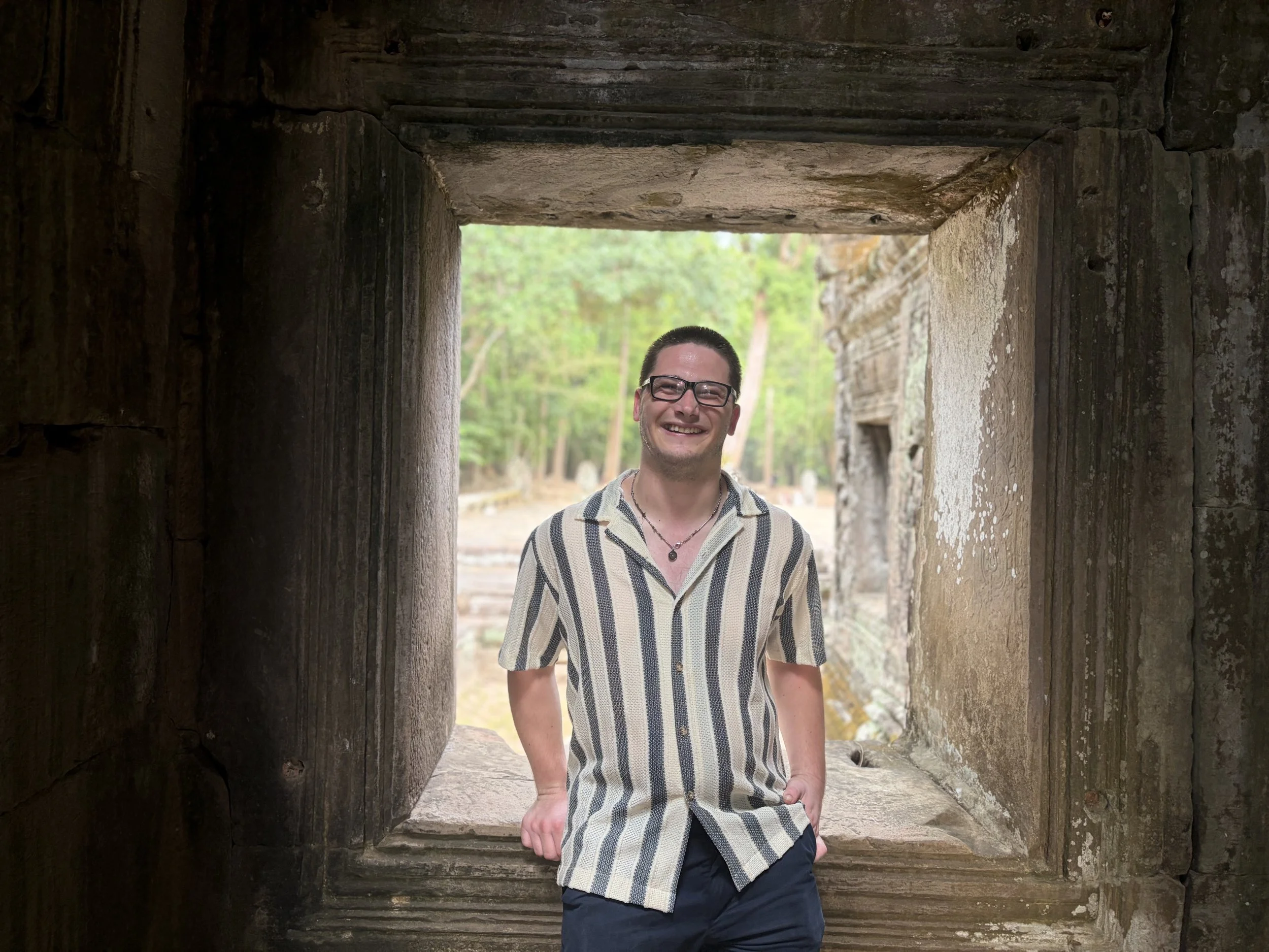 Harvey Matthewson, a young man with glasses and a striped short-sleeve shirt standing by a large wooden structure in Asia with an open rectangular window, outdoors with trees and greenery in the background.