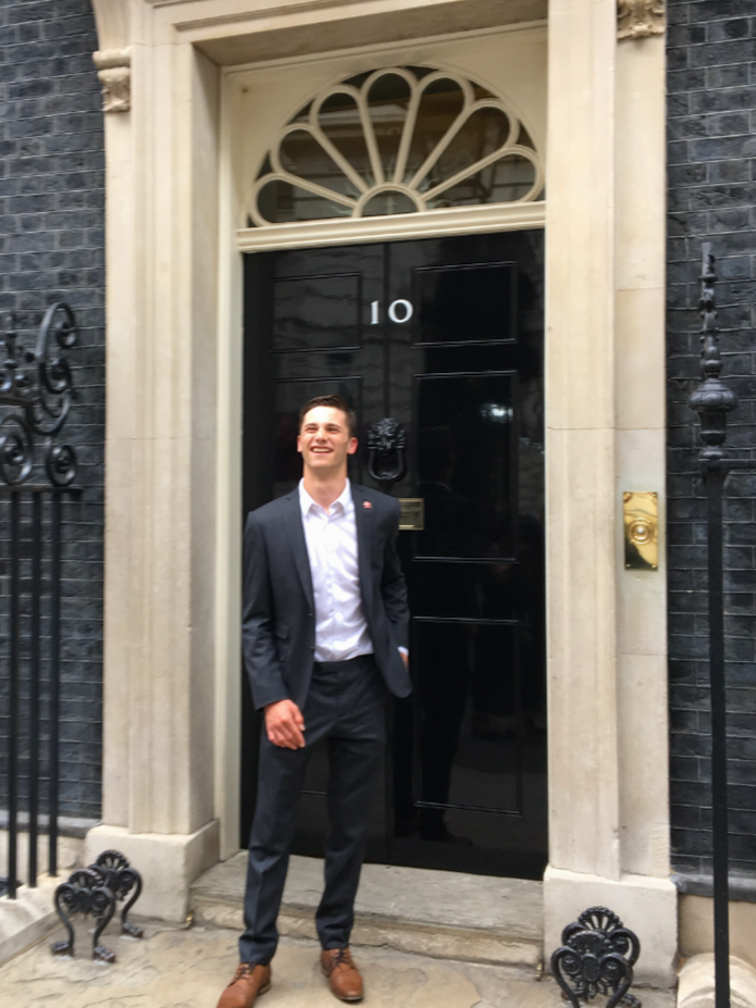 Harvey Matthewson in a suit standing in front of the black door of 10 Downing Street, representing the Kings' Trust, smiling.