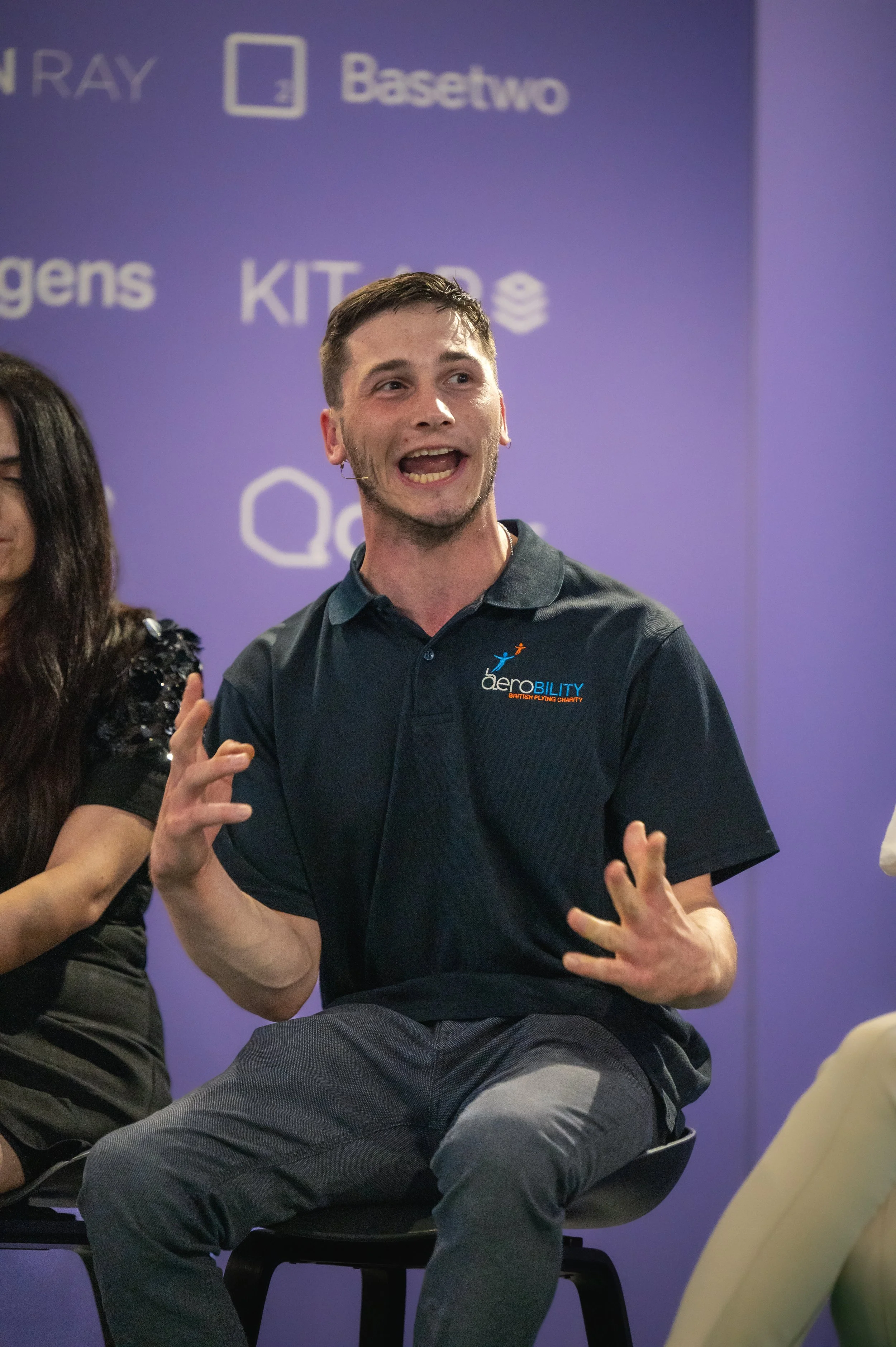 Harvey Matthewson speaking at the Farnbourgh International Airshow and gesturing with his hands, wearing a dark polo shirt with the logo 'Aerobility' on it, seated in front of a purple background with various logos and text.