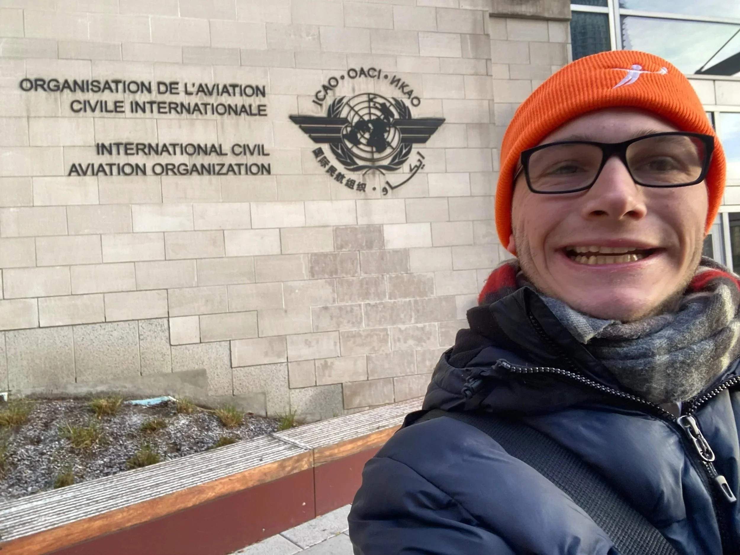 Selfie of Harvey Matthewson wearing black glasses, an orange beanie with a white logo, a black jacket, and a scarf, standing in front of a wall with the logo and name of the International Civil Aviation Organization (ICAO) in multiple languages.
