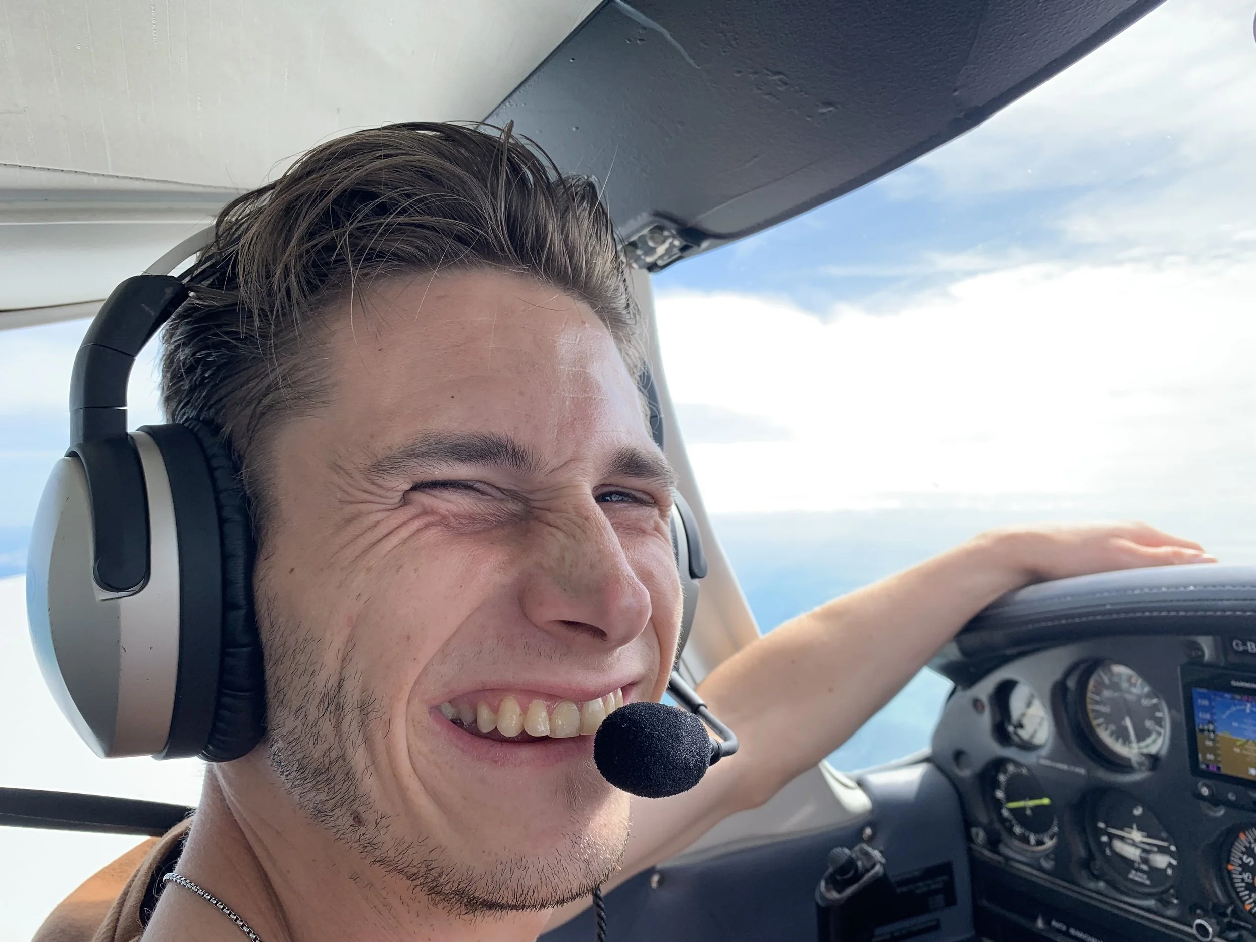 Harvey Matthewson a smiling young man wearing headphones with a microphone, sitting in the cockpit of flying aircraft, with a bright sky and clouds visible outside the window.
