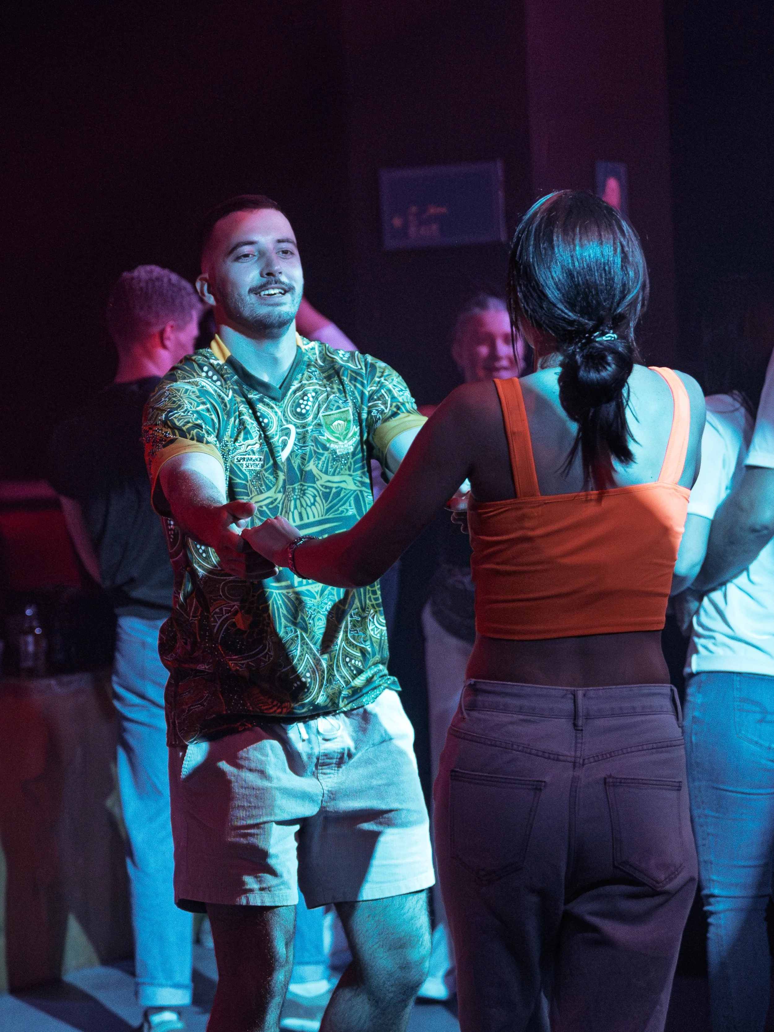 A group of people dancing together in a dimly lit nightclub or party setting with colorful lighting.
