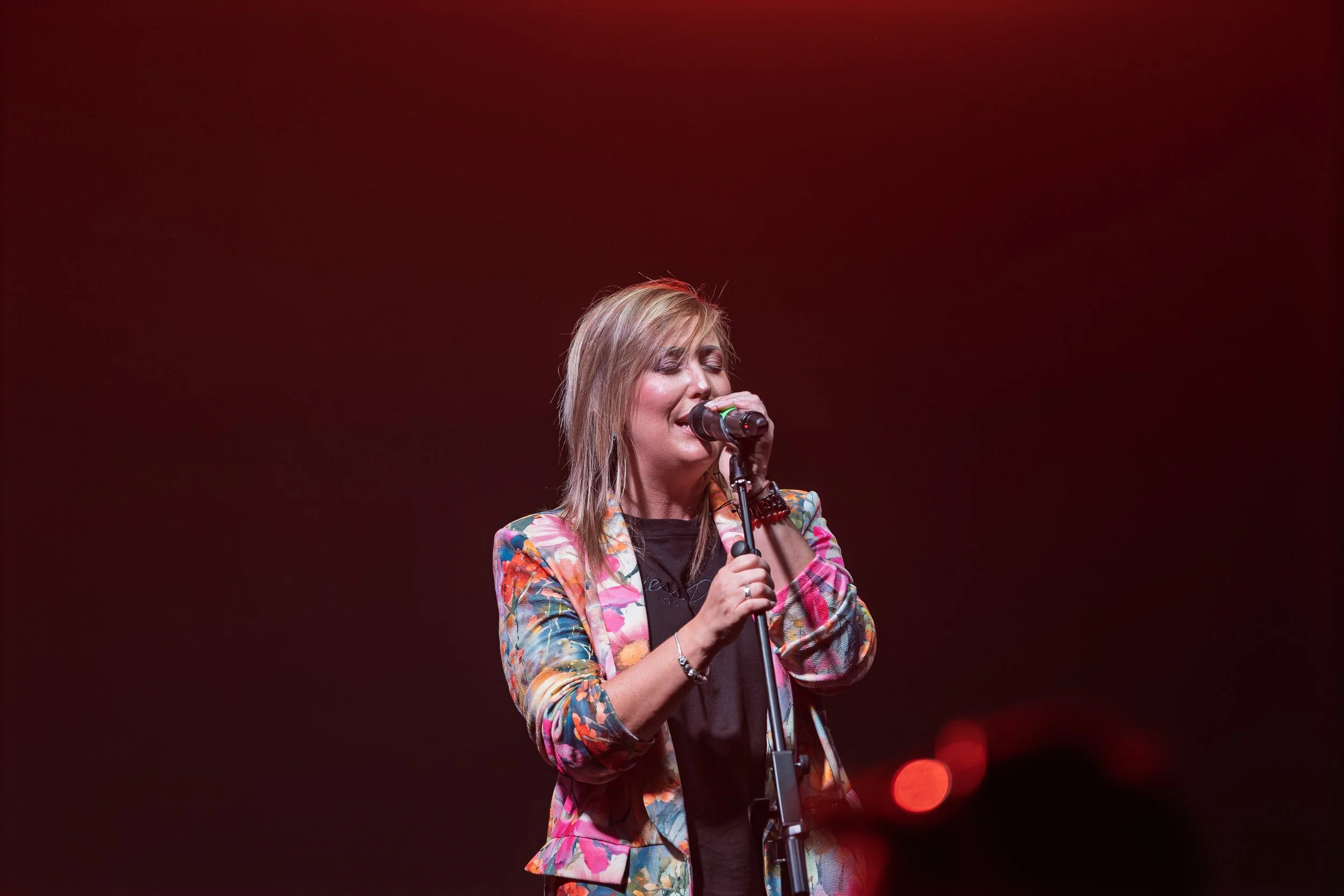 A woman singing into a microphone on stage with a dark red background.