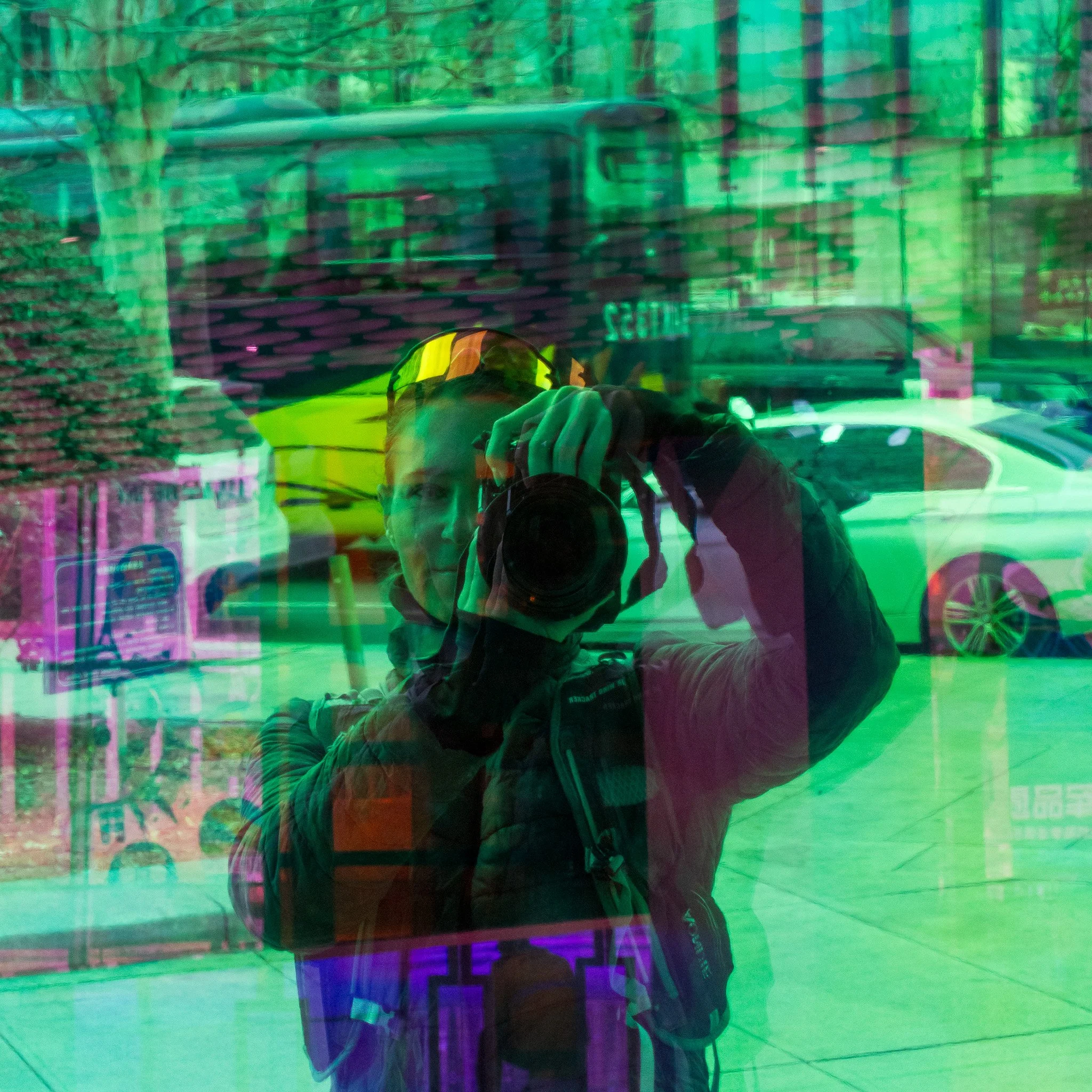 A person taking a selfie reflection on a green-tinted window or surface with a cityscape and trees in the background.