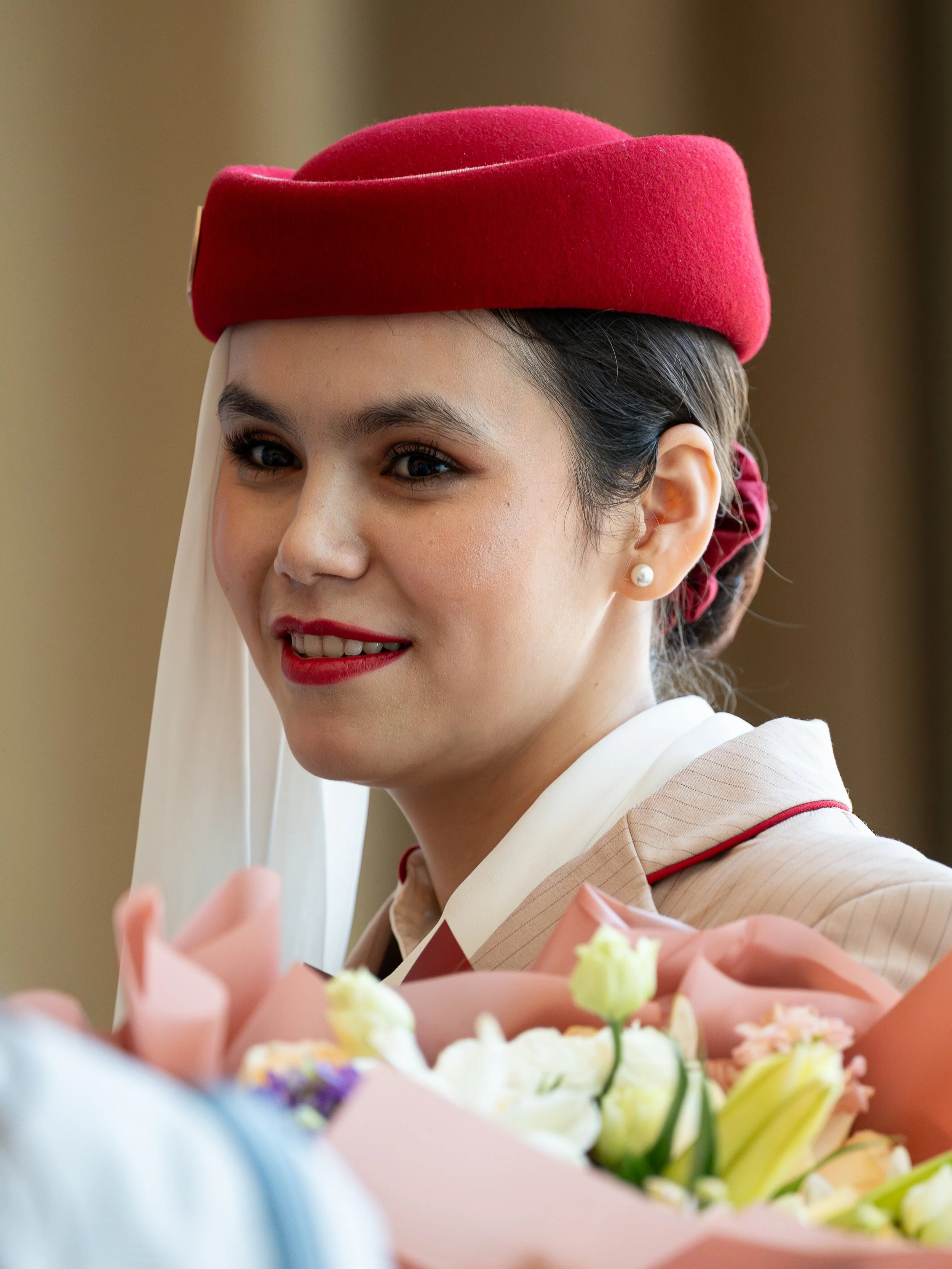 A woman wearing a red hat and red lipstick, dressed in vintage attire, is smiling and holding a bouquet of flowers.