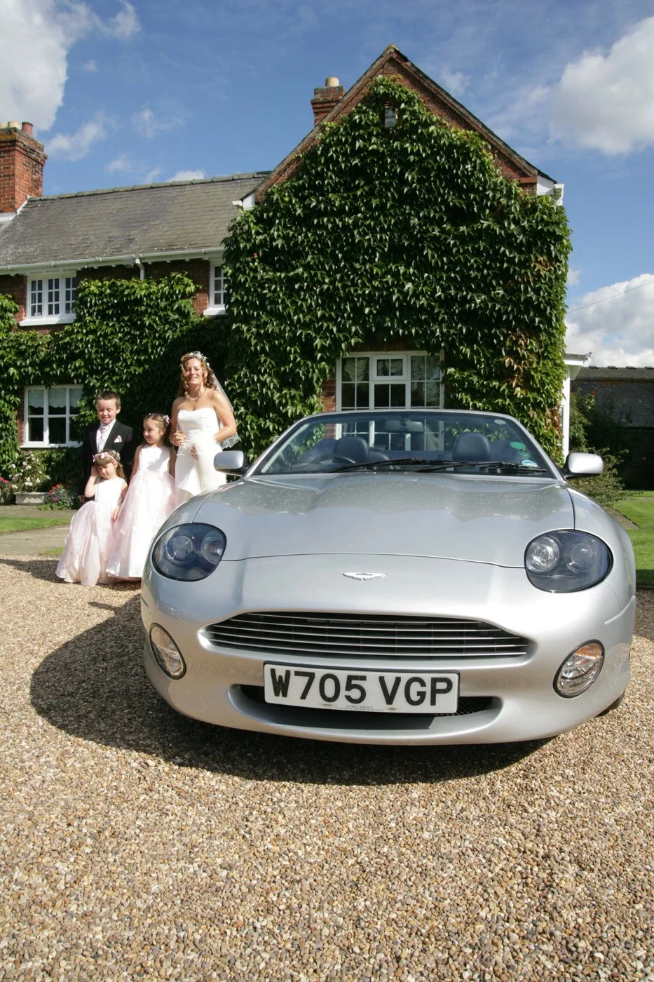 A wedding scene in front of a brick house with ivy, featuring a bride and three children in formal attire, and a silver convertible car with the license plate W705 VGP parked on gravel.