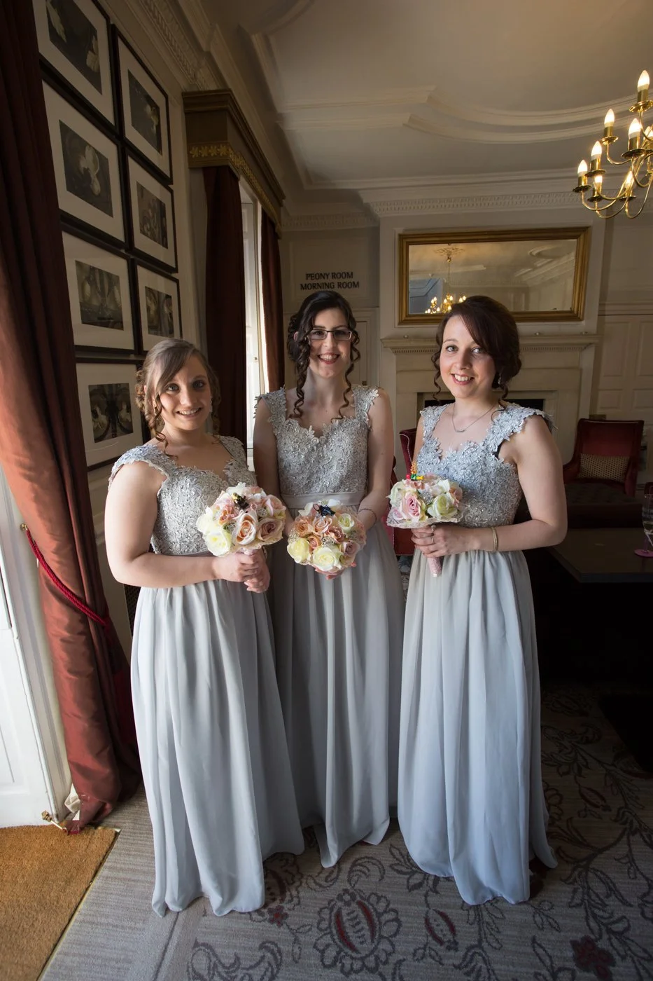 Three women in bridesmaid dresses holding bouquets, standing in a decorated room with framed photos on the wall and a mirror, smiling at the camera.