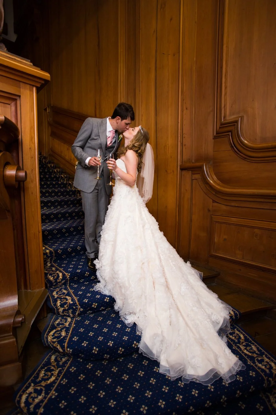 Bride and groom sharing a kiss on a staircase at their wedding reception, holding champagne glasses, in a wood-paneled room.
