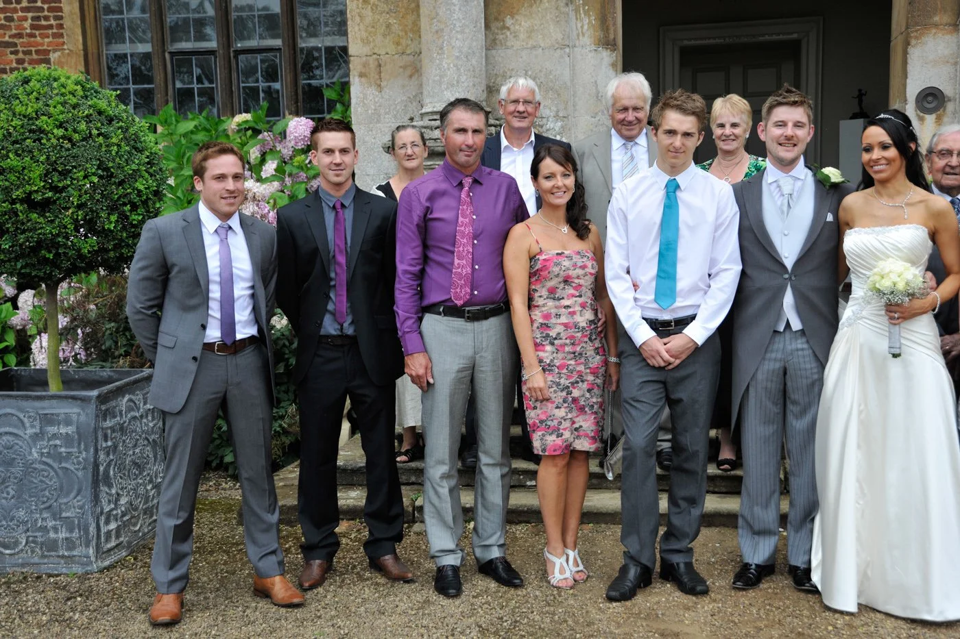 Group of people dressed for a wedding, standing outside of a historic building with stone walls and greenery.