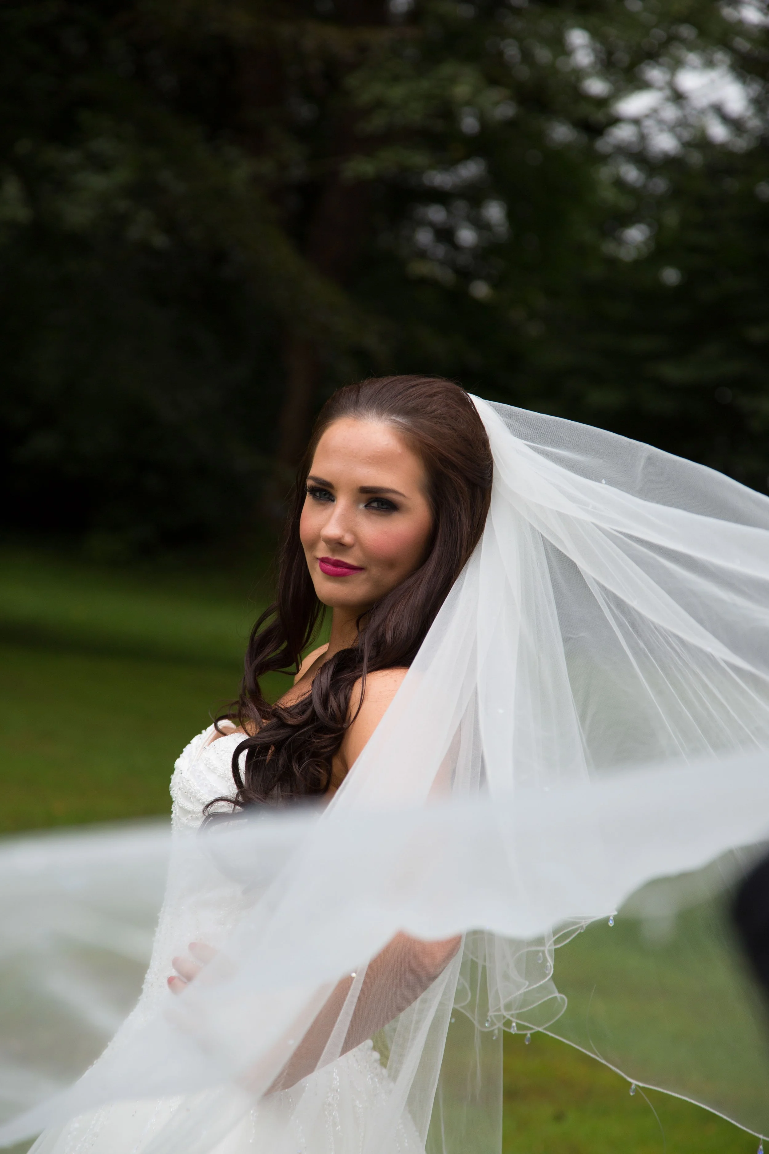 A bride with long dark hair and pink lipstick in a white wedding dress and veil standing outdoors in a green park.