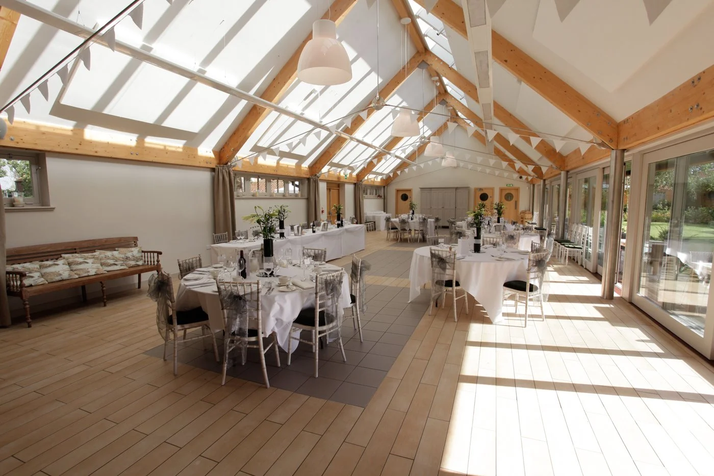 Sunlit event space with round tables covered in white tablecloths, decorated with black vases holding green flowers, set for a gathering. The room has a high, sloped ceiling with exposed wooden beams, large windows, and sliding glass doors allowing s