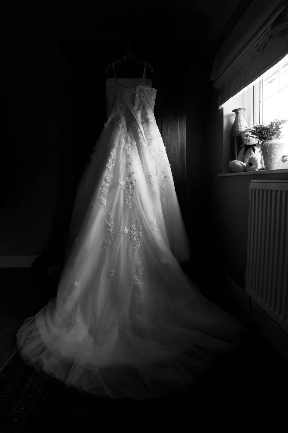 A wedding dress hanging on a hanger in a dimly lit room, with a window letting in natural light and a vase with flowers on the windowsill.