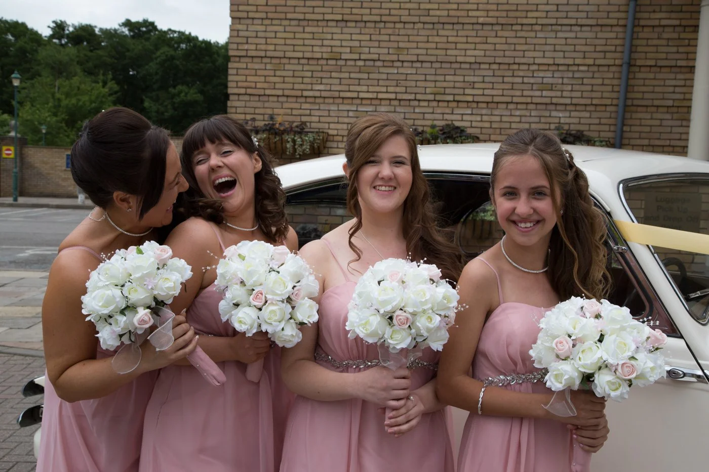Four women in pink bridesmaid dresses holding white bouquets standing next to a white vintage car outdoors, smiling and celebrating.