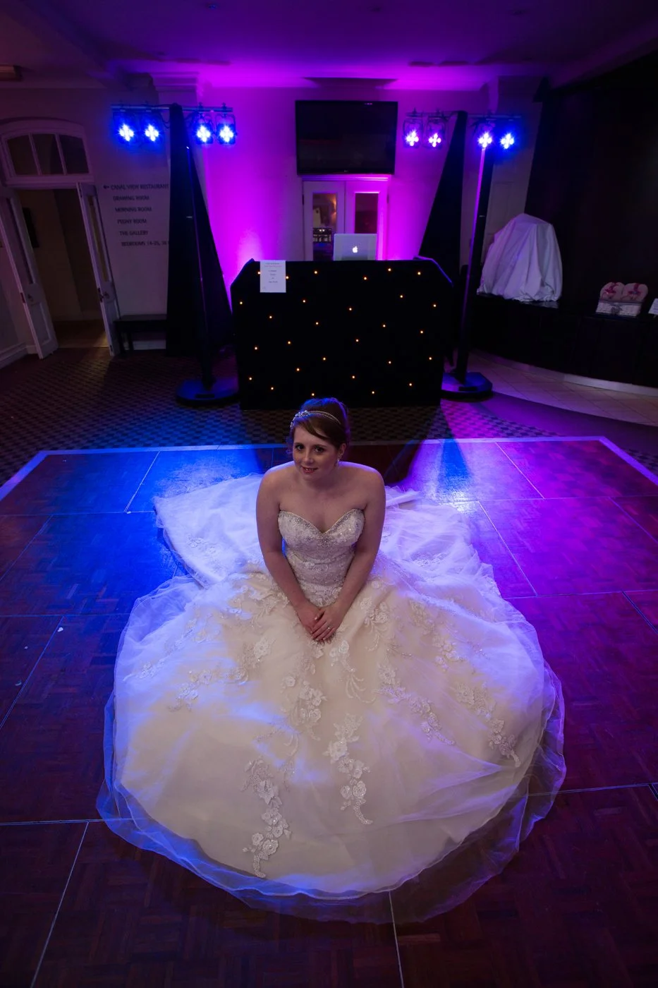 Bride in a strapless wedding gown sitting on the dance floor in a dimly lit reception hall with purple and blue lighting and a DJ table in the background.