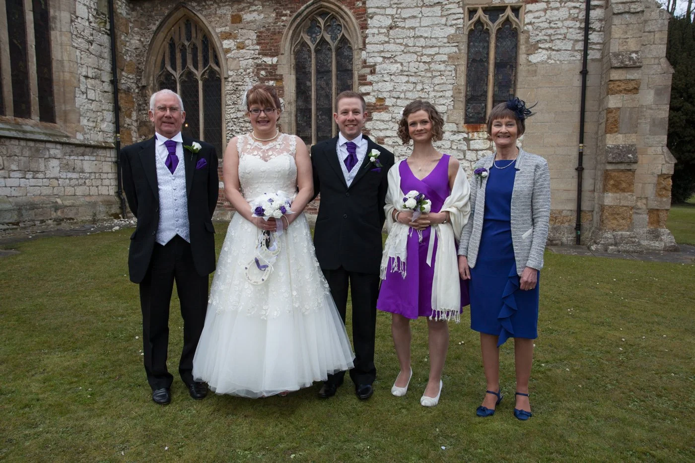 Group of six people, including a bride and groom, standing outside a stone church. The bride is wearing a white gown with lace details, holding a bouquet. The groom is in a black suit with a purple tie and boutonniere. Two women are dressed in purple