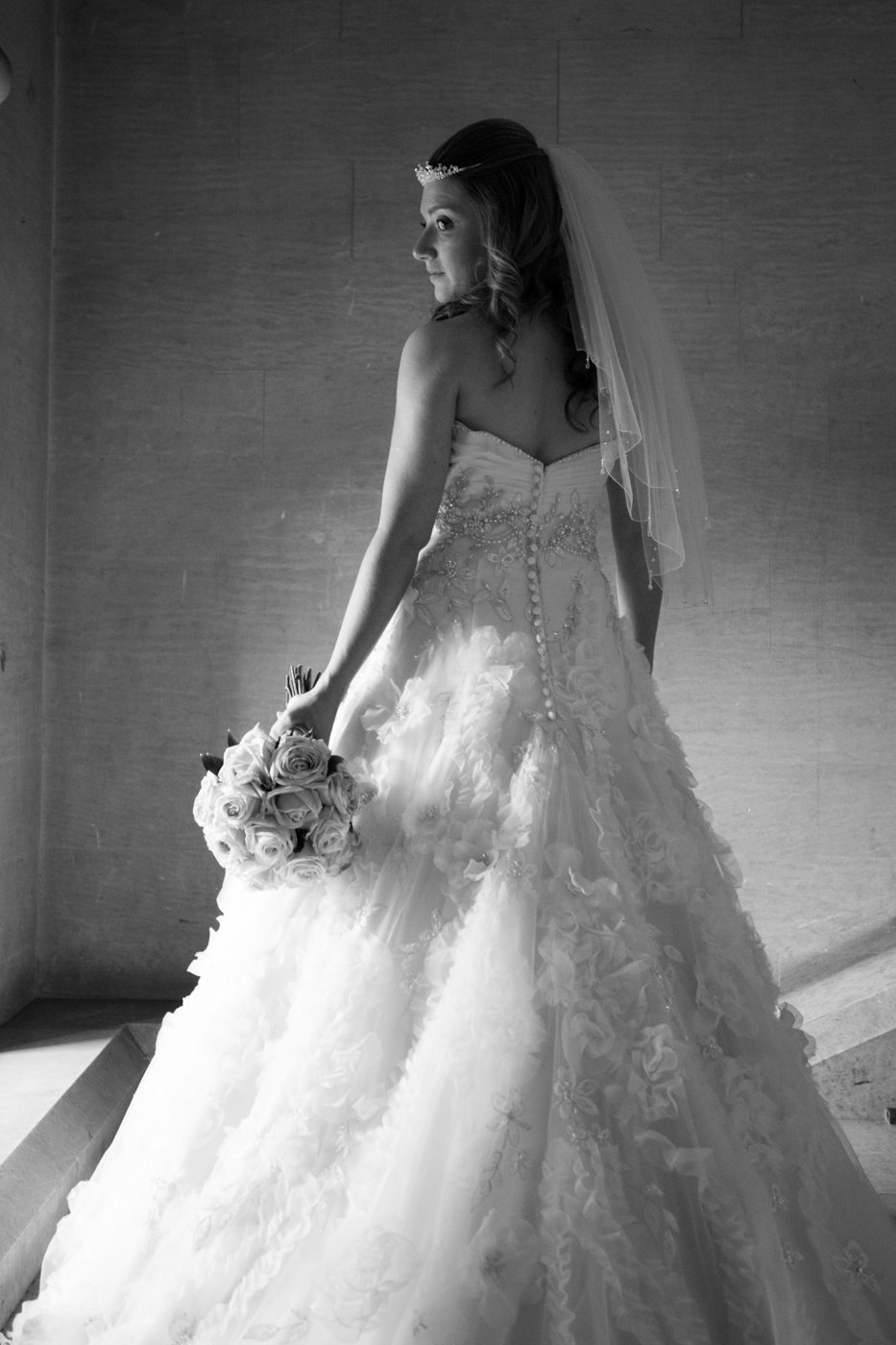 Black and white photograph of a bride in a wedding gown holding a bouquet of roses, standing against a plain wall.