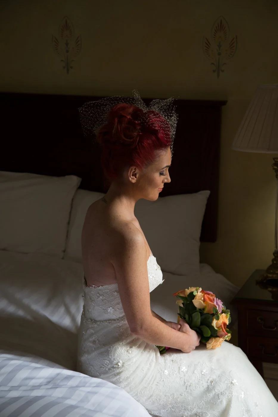 A bride with red hair in a white wedding dress sitting on a bed, holding a bouquet of roses, with her eyes closed and head tilted slightly downward.