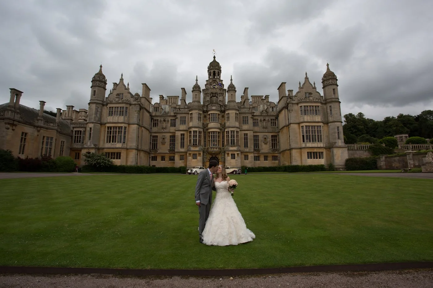 A bride and groom in wedding attire sharing a kiss in front of a large, historic castle on a cloudy day.