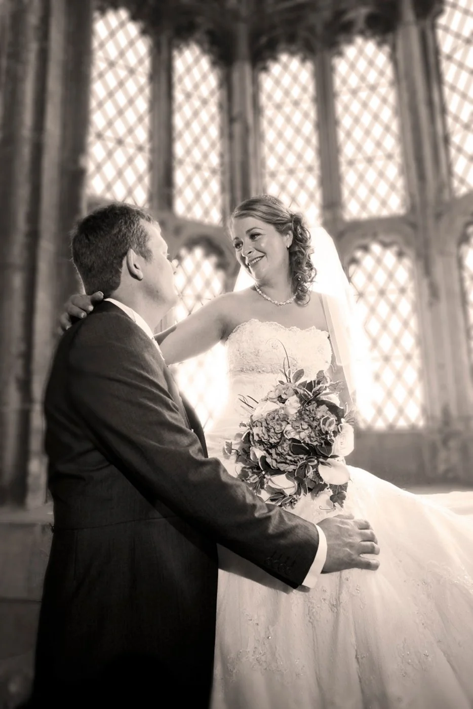 A bride and groom on their wedding day inside a church, with the bride holding a bouquet of flowers, smiling at each other.