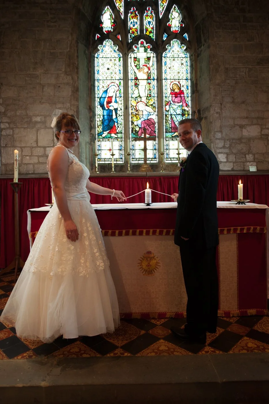 Bride and groom holding a candle lighter in a church during their wedding ceremony, standing in front of an altar with a stained glass window depicting a religious scene.