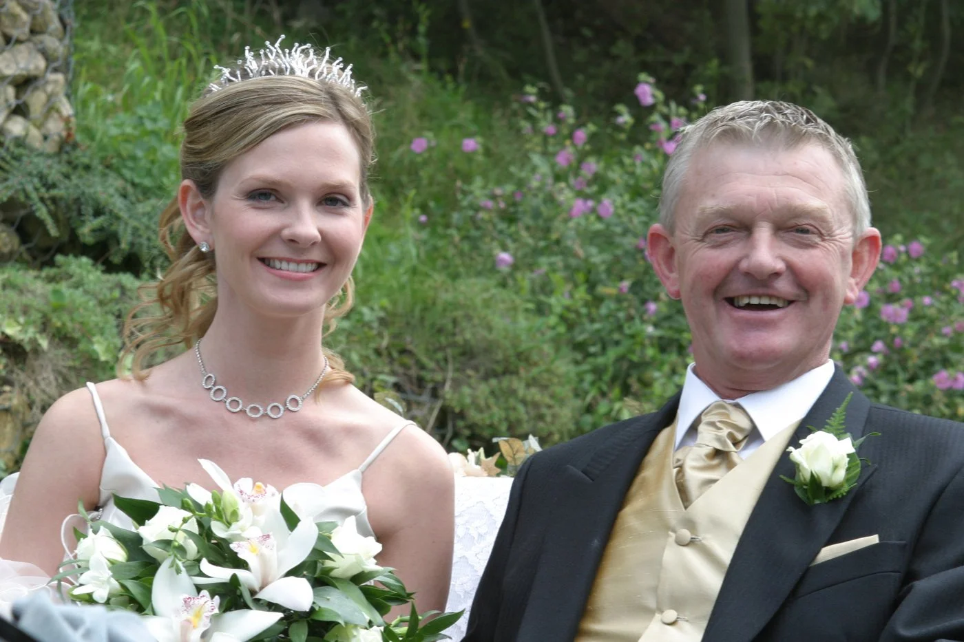 A bride and an older man, possibly her father, smiling at a wedding ceremony outdoors, with flowers and greenery in the background.