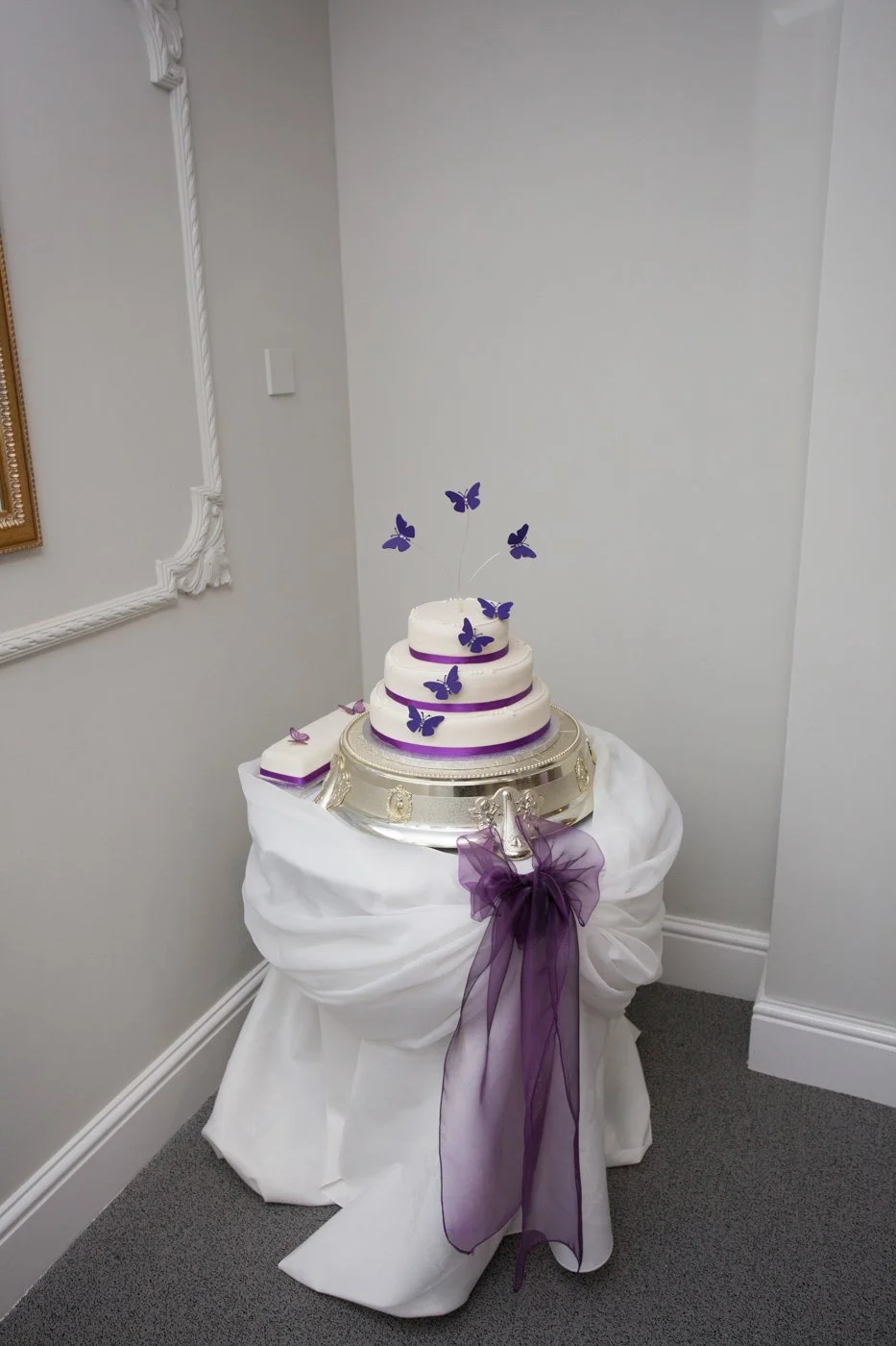 A multi-tiered white cake decorated with purple ribbons and purple butterfly toppers, displayed on a silver cake stand draped with white cloth and purple sheer ribbon, in a decorated corner of a room.