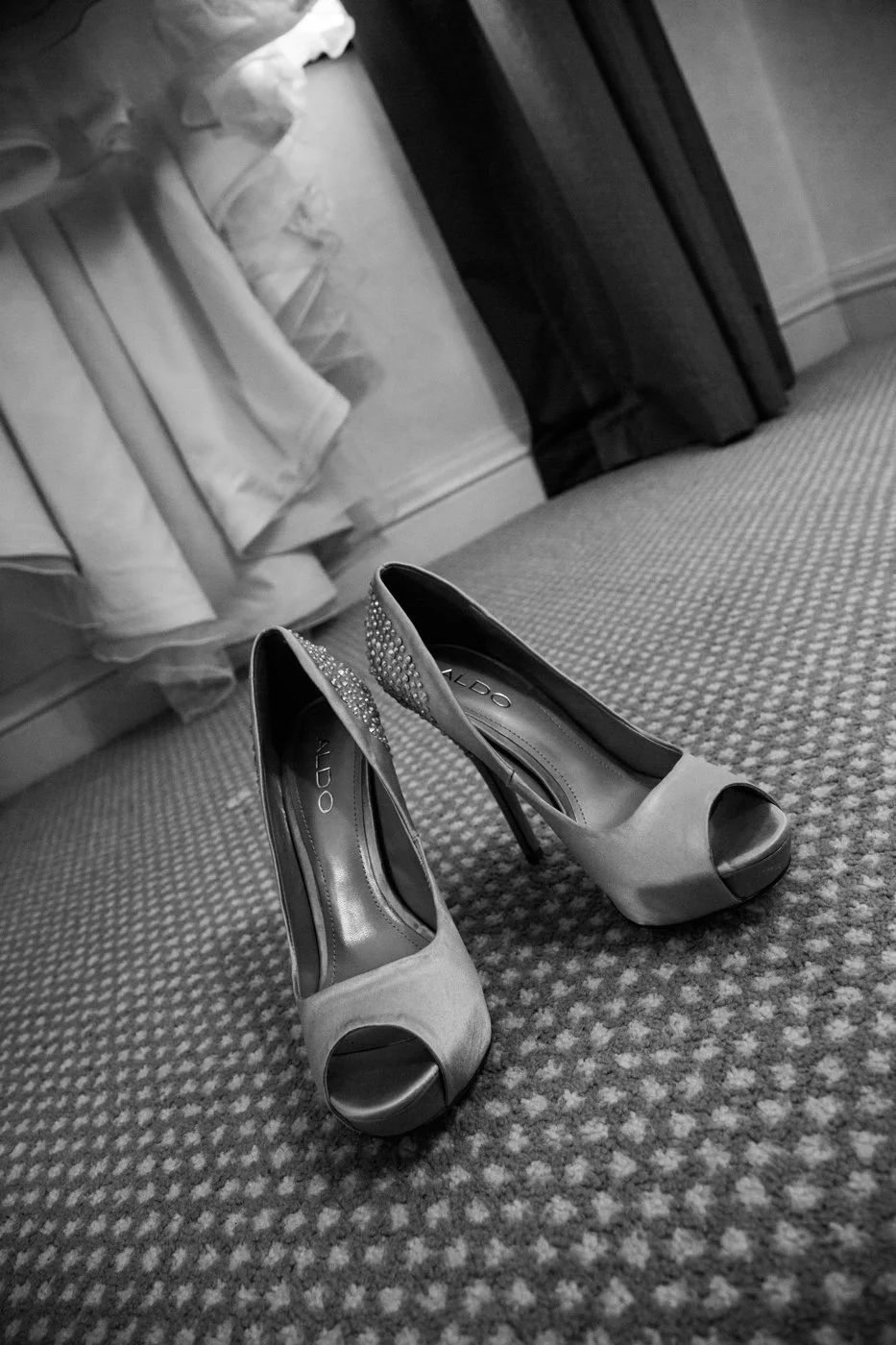 A pair of high-heeled women's shoes with peep toes, decorated with rhinestones on the back, placed on a patterned carpeted floor near a bed with ruffled bedding.