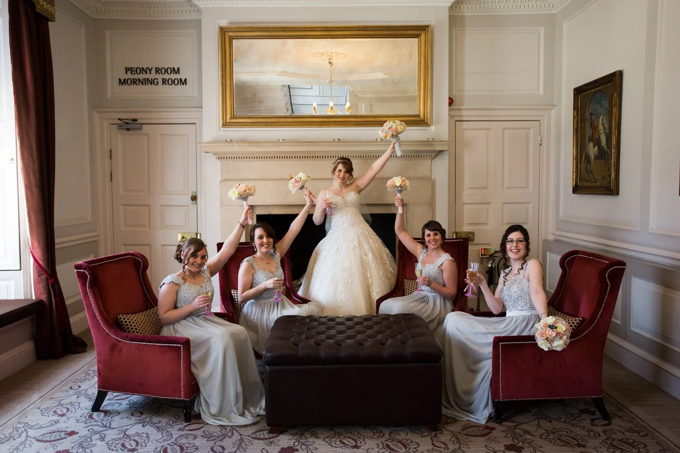 A bride in a wedding gown and five bridesmaids in matching dresses celebrating in a decorated room. They are sitting and standing on red upholstered chairs and holding bouquets and glasses, with the bride raising her arm. A mirror reflects a chandeli