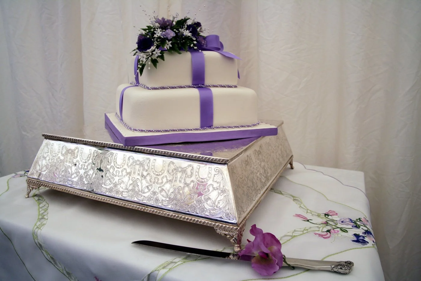 A two-tiered wedding cake with purple ribbons and a floral decoration on top, placed on a decorative silver cake stand. The tablecloth has floral embroidery, and a pink flower is next to a cake serving utensil.