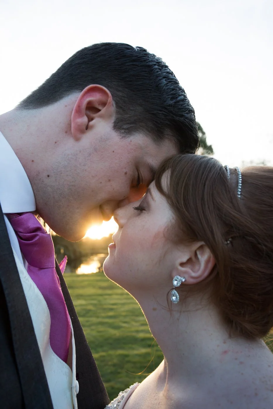 A close-up photo of a bride and groom with foreheads touching during sunset, outdoors in a park.
