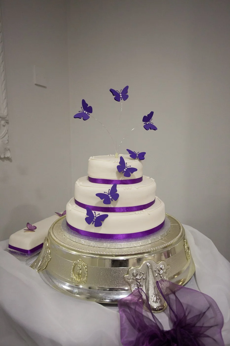 A three-tier white cake decorated with purple ribbons and purple butterfly toppers, placed on a silver cake stand with purple tulle decoration in front.