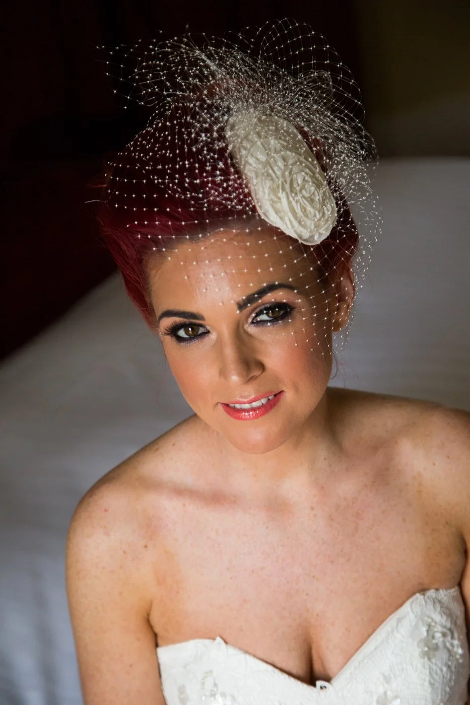 A woman with red hair and freckles wearing a white strapless dress, smiling with one eye winking, decorated with a digital floral headpiece and a netted veil.