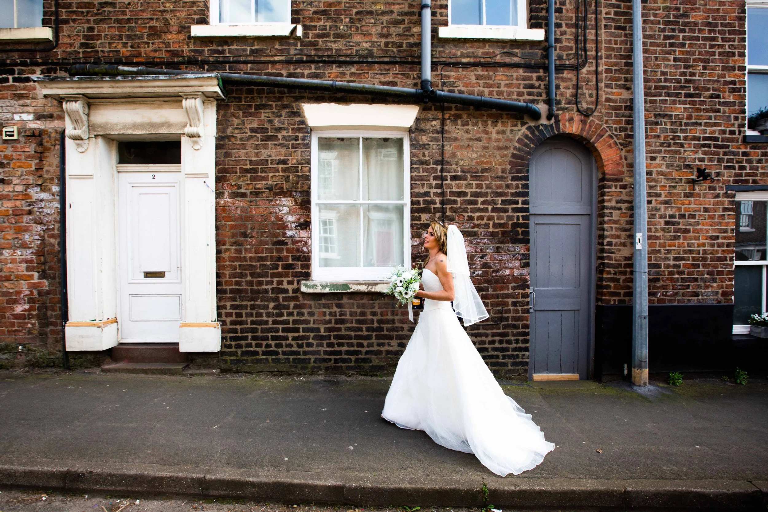 A woman in a white wedding dress holding a bouquet of white flowers, standing on a sidewalk in front of a brick building with white and gray doors and windows.