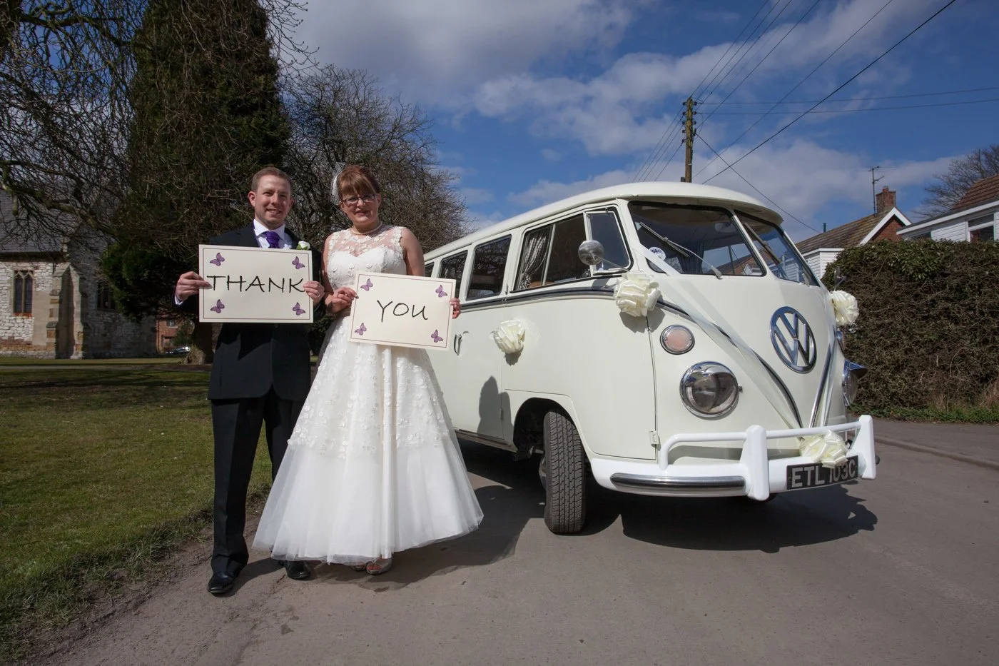 A newlywed couple standing outdoors near a vintage white Volkswagen camper van, holding signs that say 'Thank' and 'You' decorated with purple butterflies, on a sunny day with partly cloudy skies and houses in the background.