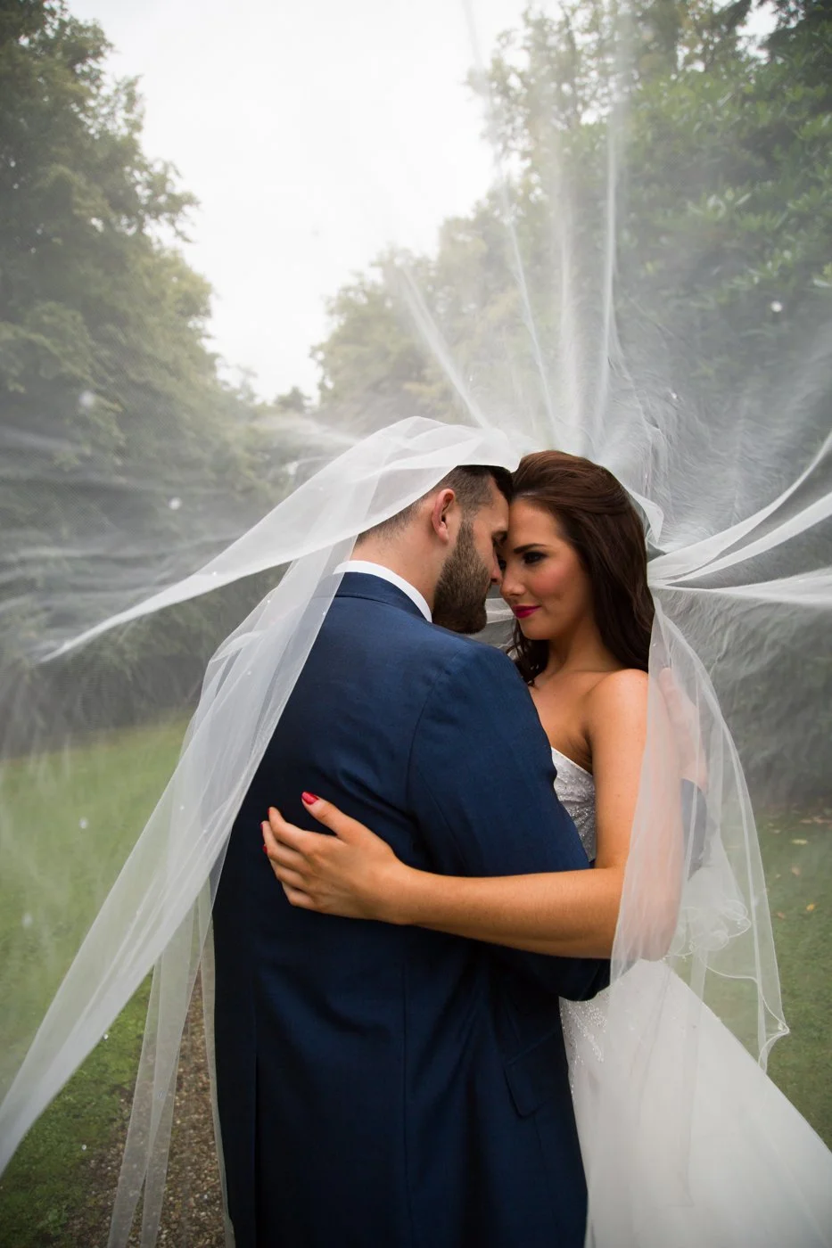 A bride and groom embracing outdoors, with the bride's veil flowing around them, under a misty sky and green trees.