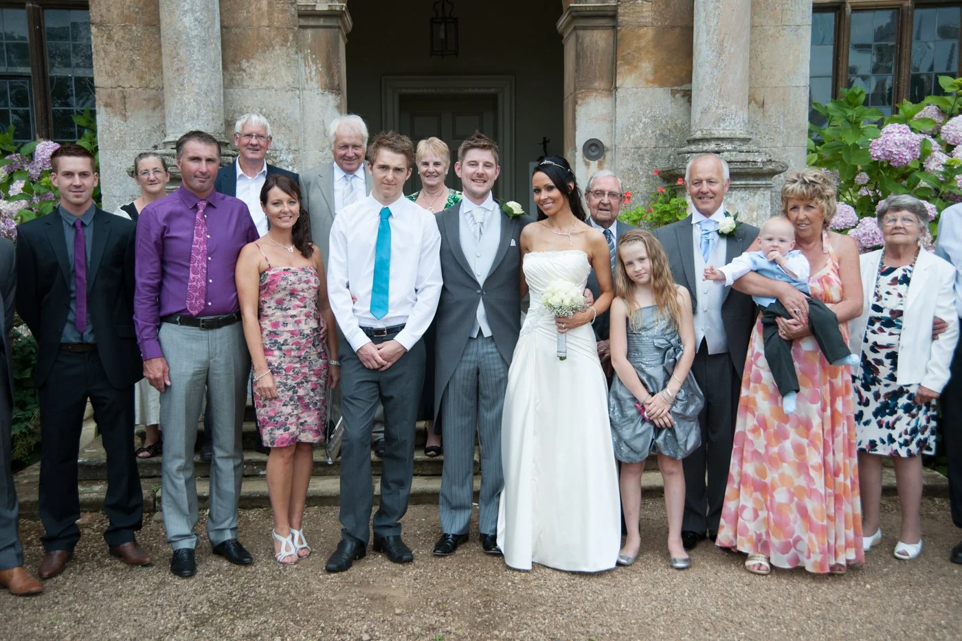 A large wedding group photo outside a stone building, with bride and groom in the center, surrounded by family and friends.
