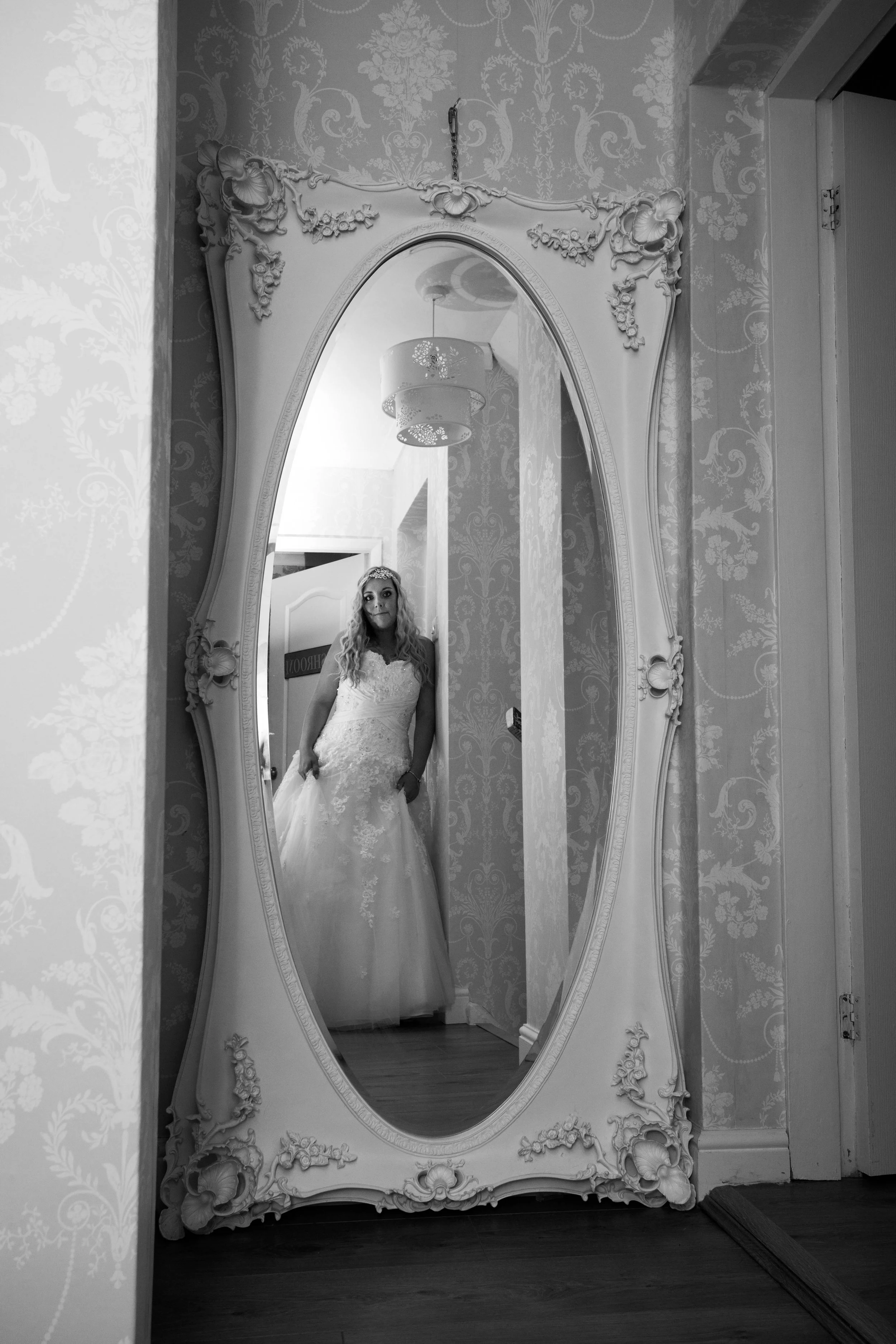 A woman in a wedding dress is taking a selfie in a mirror with ornate floral carvings, reflected in the mirror, and floral wallpaper in the background.