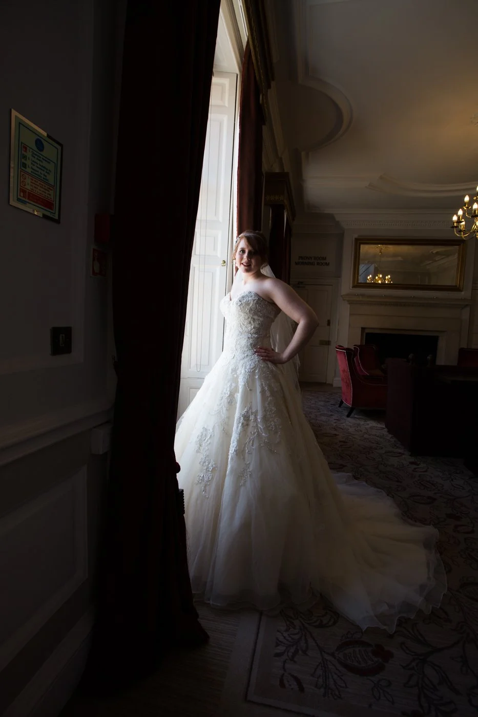 Bride in a white lace wedding gown standing in a dimly lit room near a large window with dark curtains, elegant interior with a fireplace and chandelier.