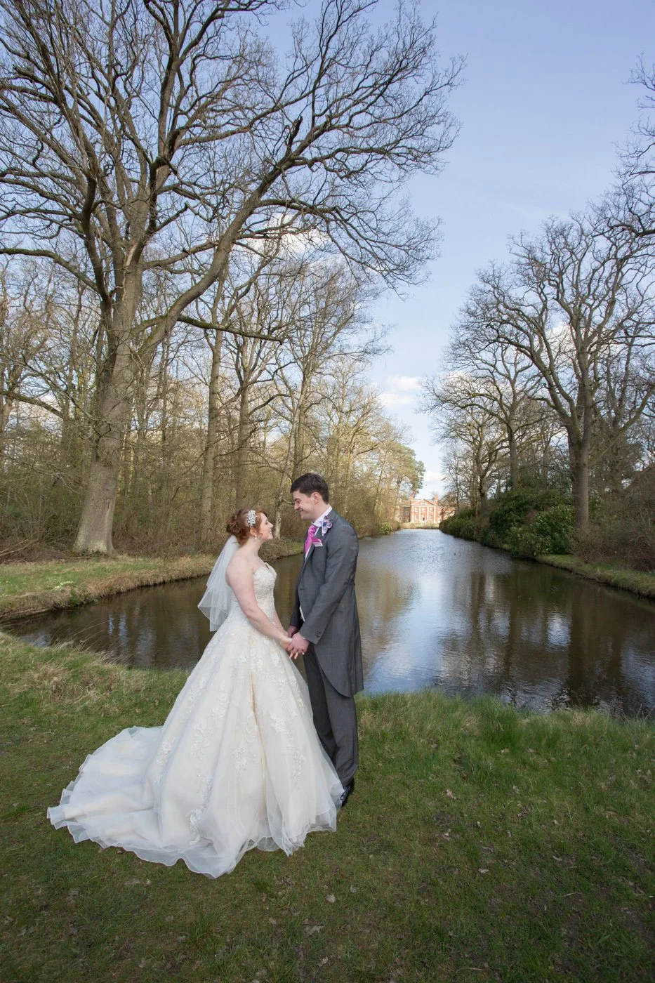 A bride and groom standing by a pond in a park, holding hands and looking at each other, with trees and a building in the background.